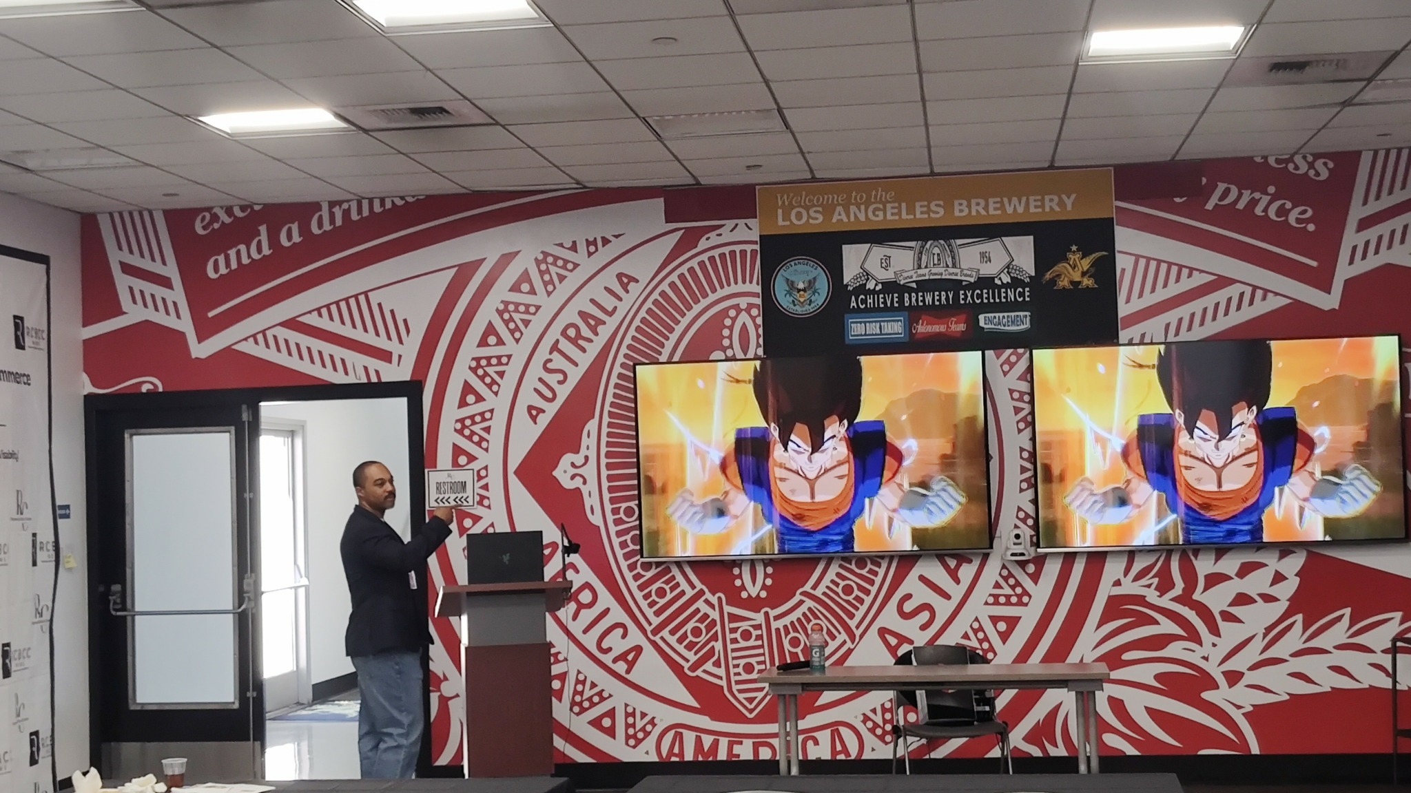 Man standing at podium in front of large screens displaying anime characters, with a red and white patterned wall behind.