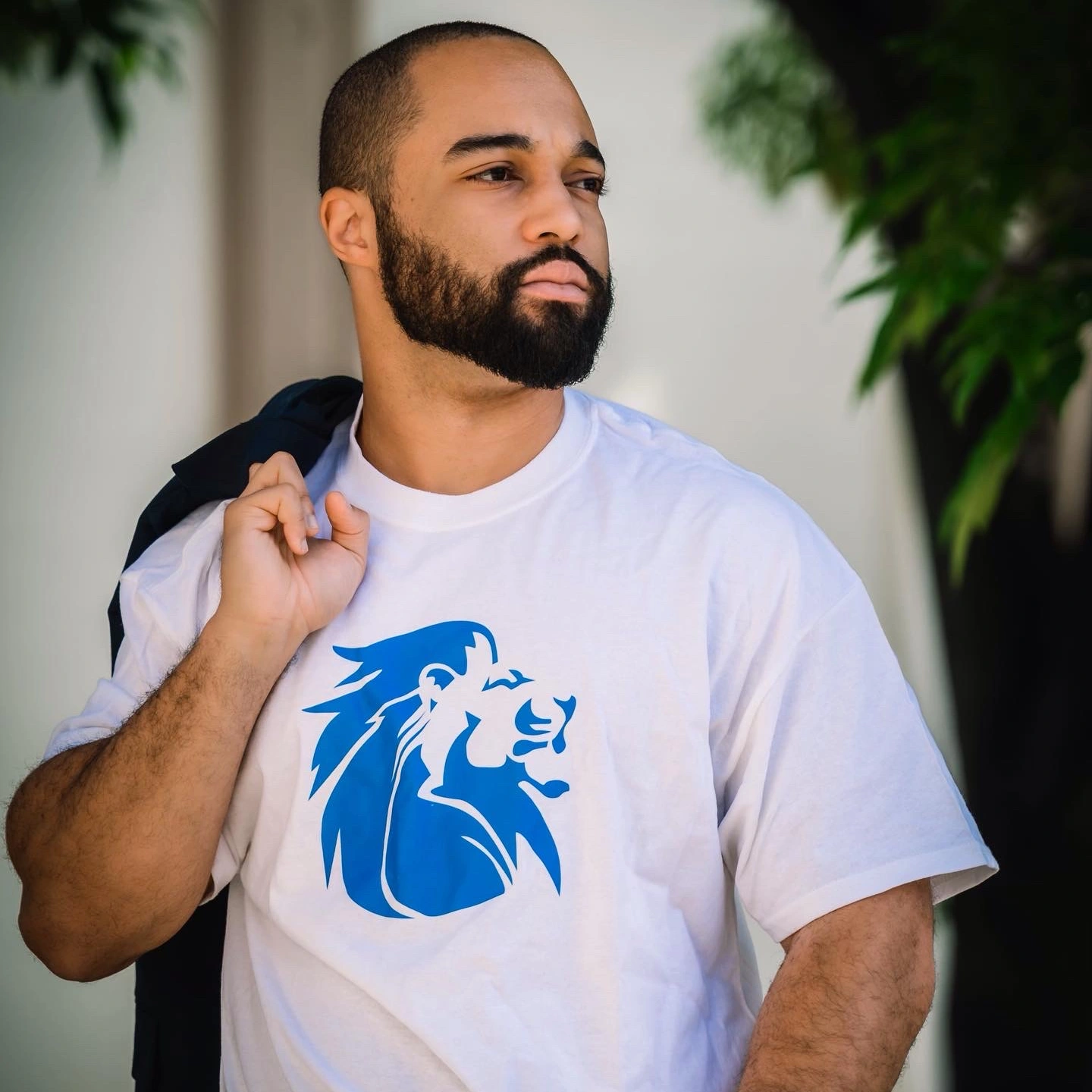 Man with beard and short hair wearing a white t-shirt with a blue lion logo, holding a jacket over his shoulder.