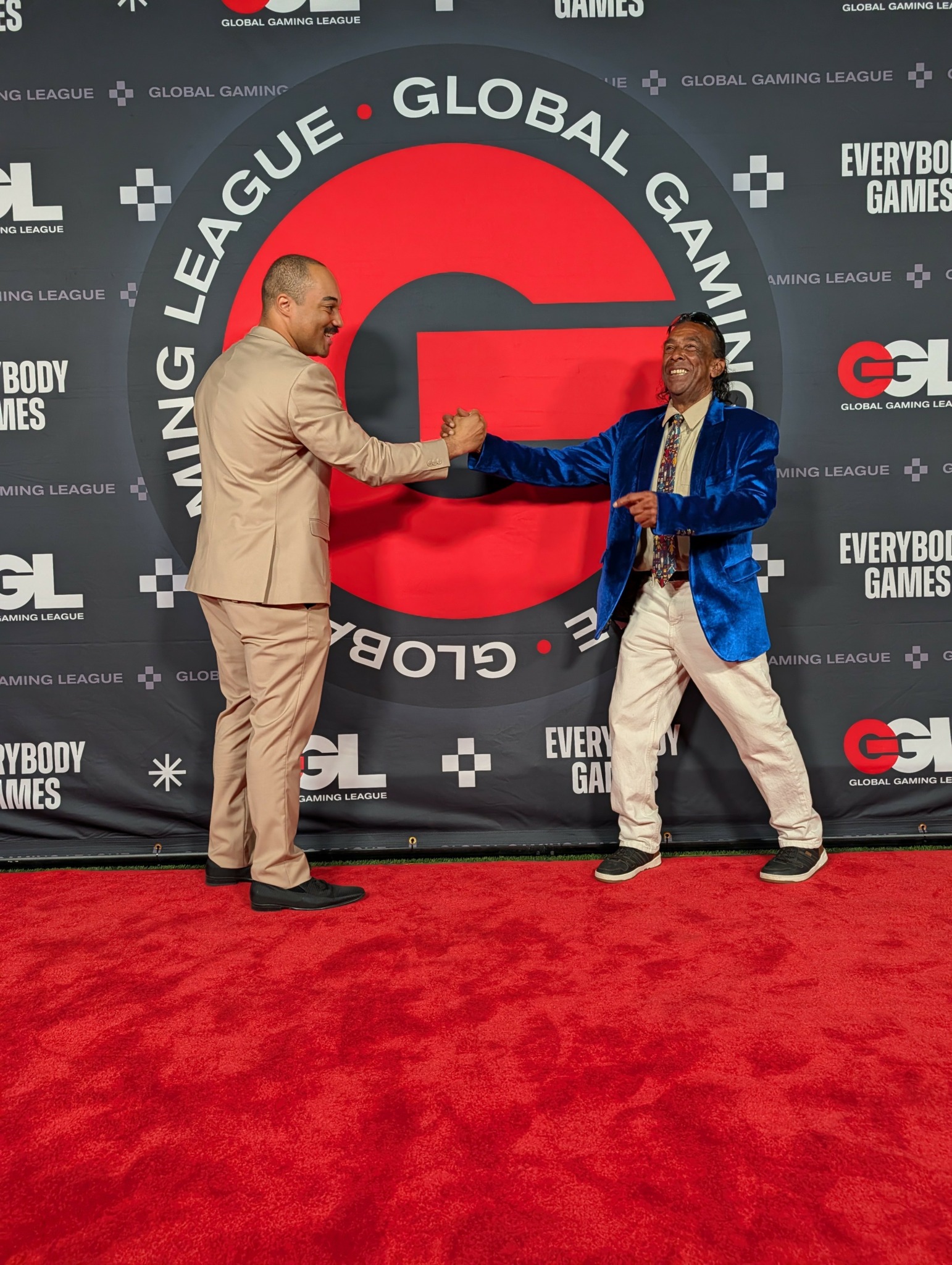 Two men shake hands on a red carpet in front of a backdrop with logos, one in a beige suit, the other in a blue blazer.