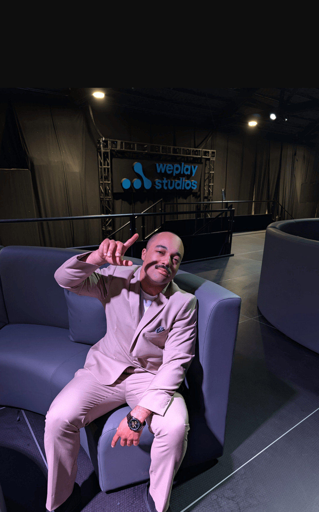Person sitting on a sofa, smiling, making a hand gesture, in a studio with a sign that reads 'wenly studios'.