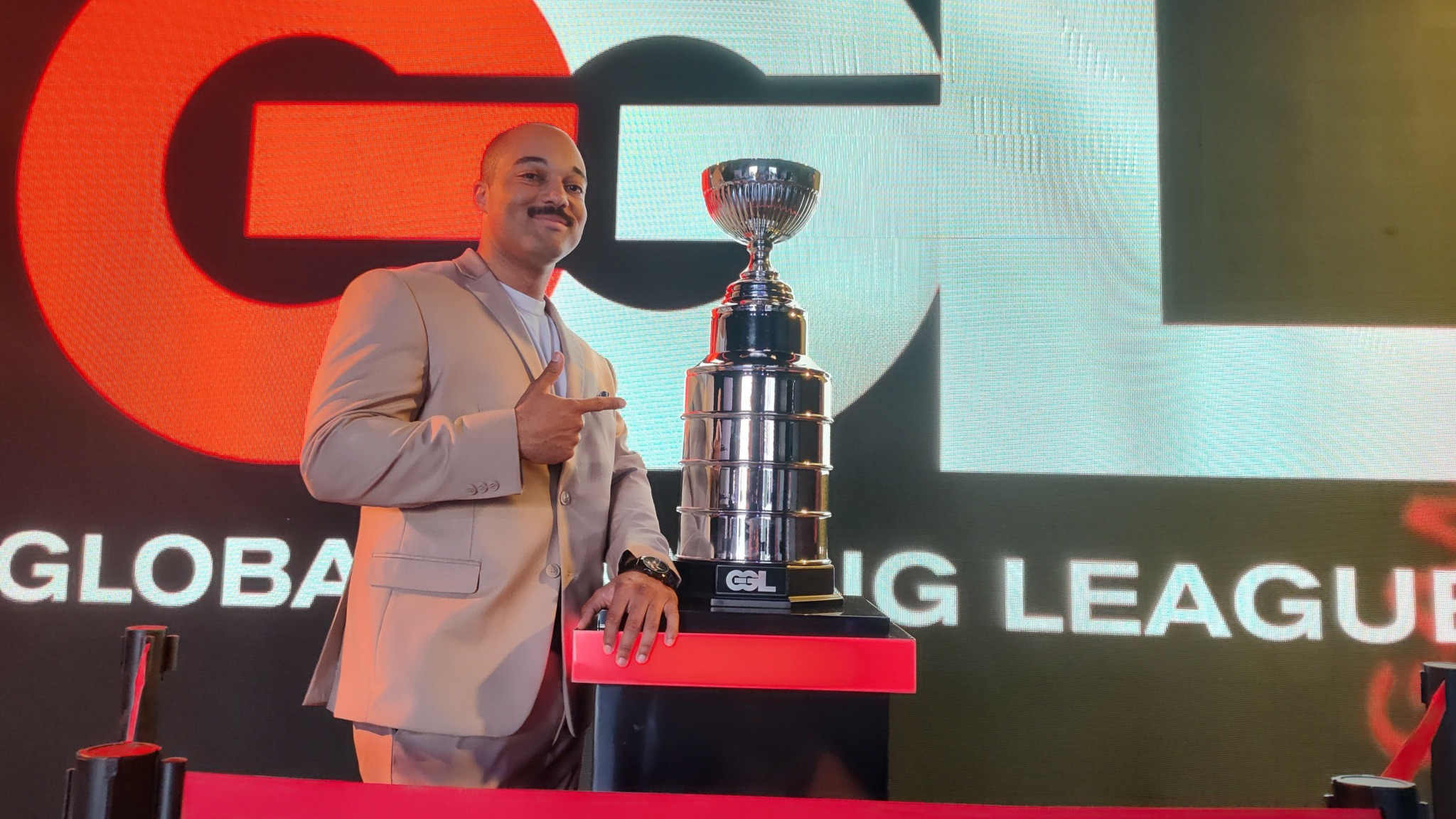 Person in suit pointing at a large trophy on a stage with a screen behind displaying a logo.