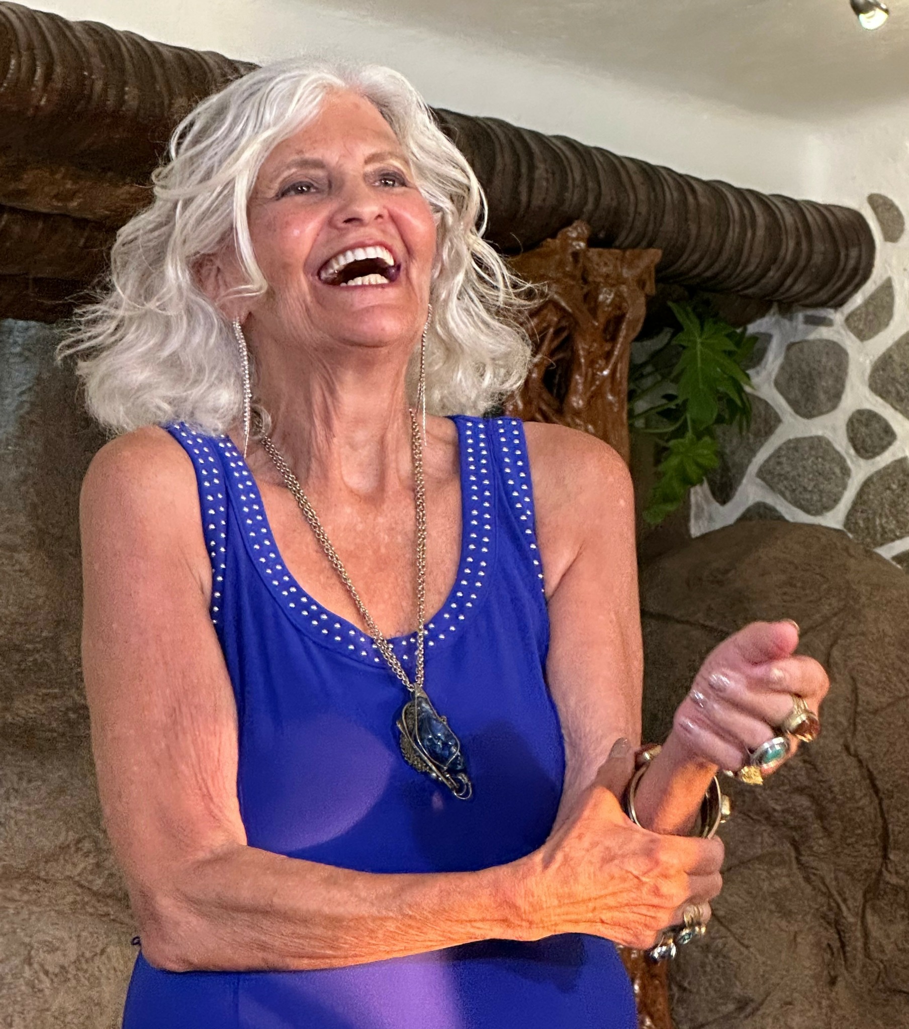 Older woman with curly white hair laughing, wearing a blue sleeveless top and jewelry, sitting indoors.