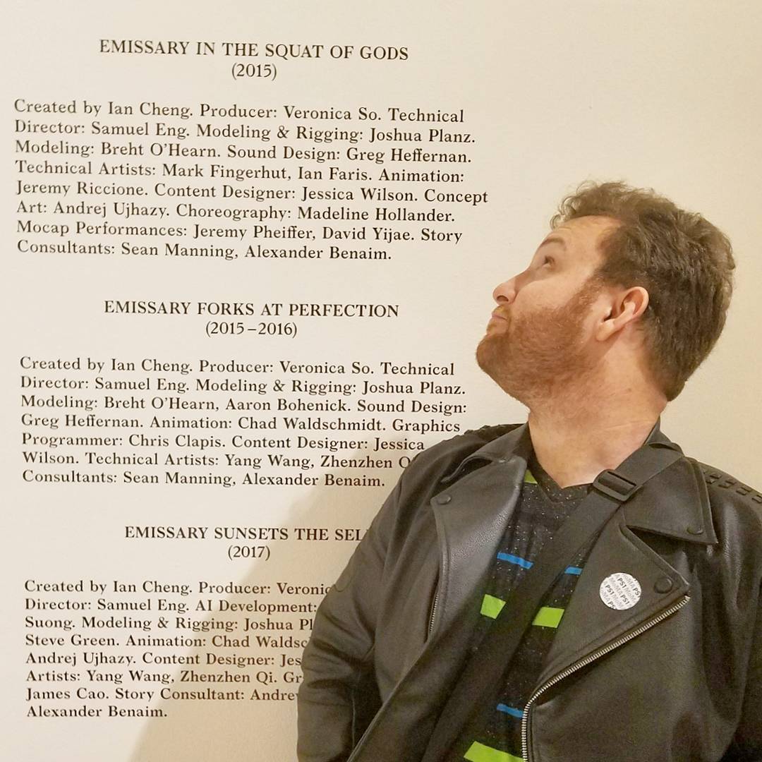Person with short hair wearing a black jacket with green accents, standing in front of a wall with text.