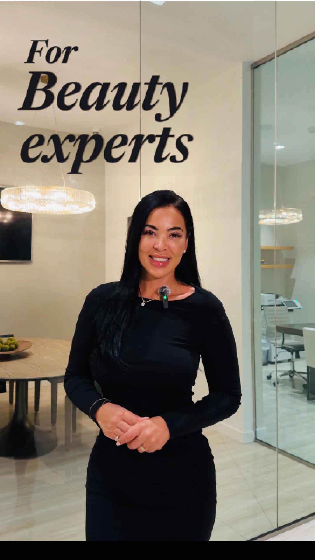 Woman with long dark hair smiling in a modern office space with glass walls and hanging lights.