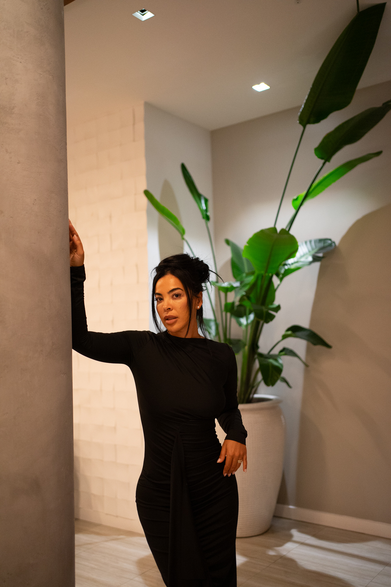 Woman in black outfit standing indoors next to large potted plant with broad leaves.