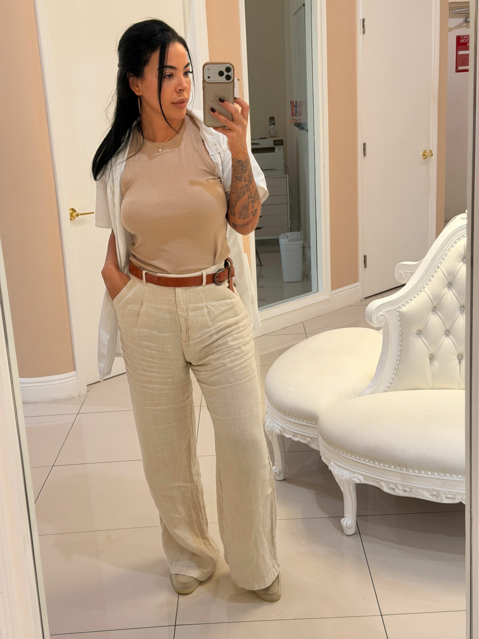 Woman in beige outfit taking mirror selfie in a room with white furniture and tiled floor.