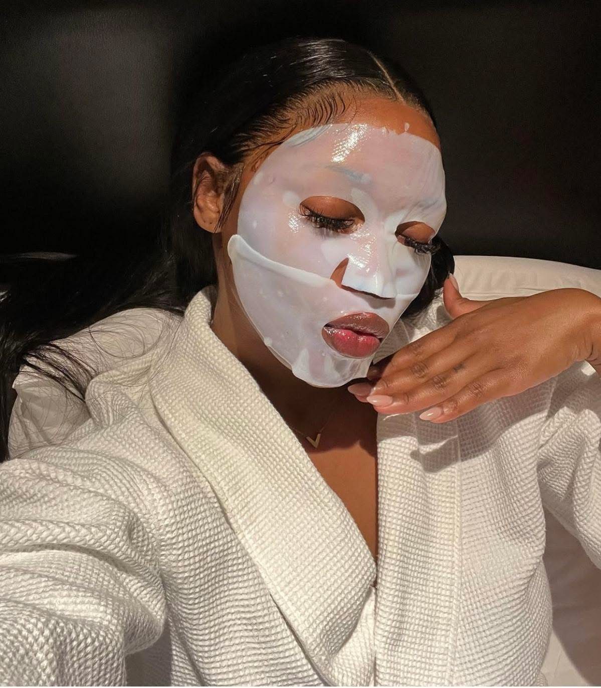 Woman with a facial mask, eyes closed, wearing a white robe, resting her hand near her face.