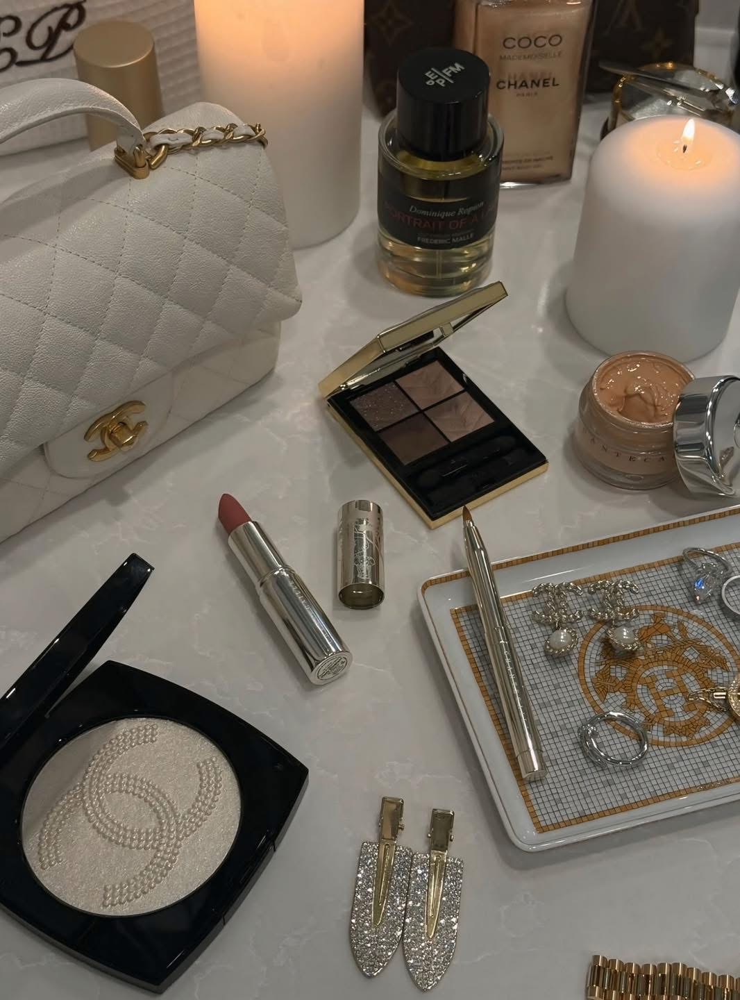 Makeup and accessories on a white surface, including a quilted white purse, eyeshadow palette, lipstick, jewelry, and a candle.