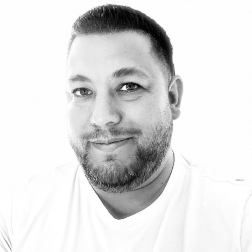 Man with short hair and beard smiling, wearing a white shirt, facing camera.