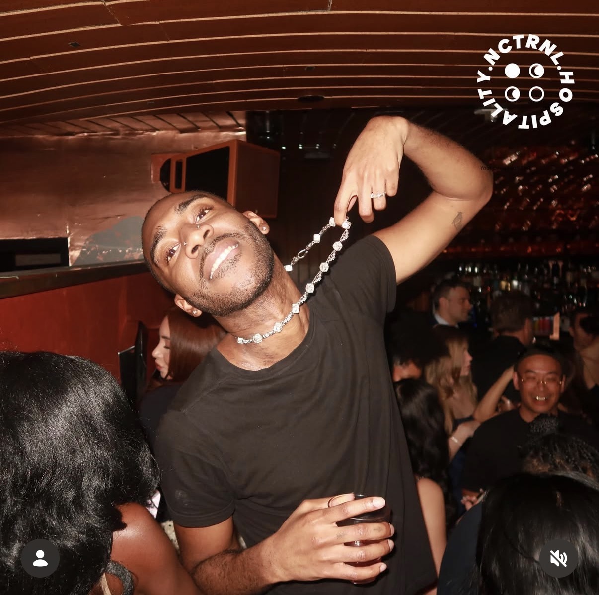 Man smiling and holding a chain with a drink in a crowded indoor setting.