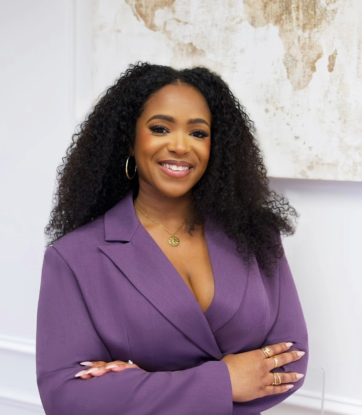 Woman with curly hair smiling, wearing a purple blazer and gold jewelry, standing indoors against a textured wall.