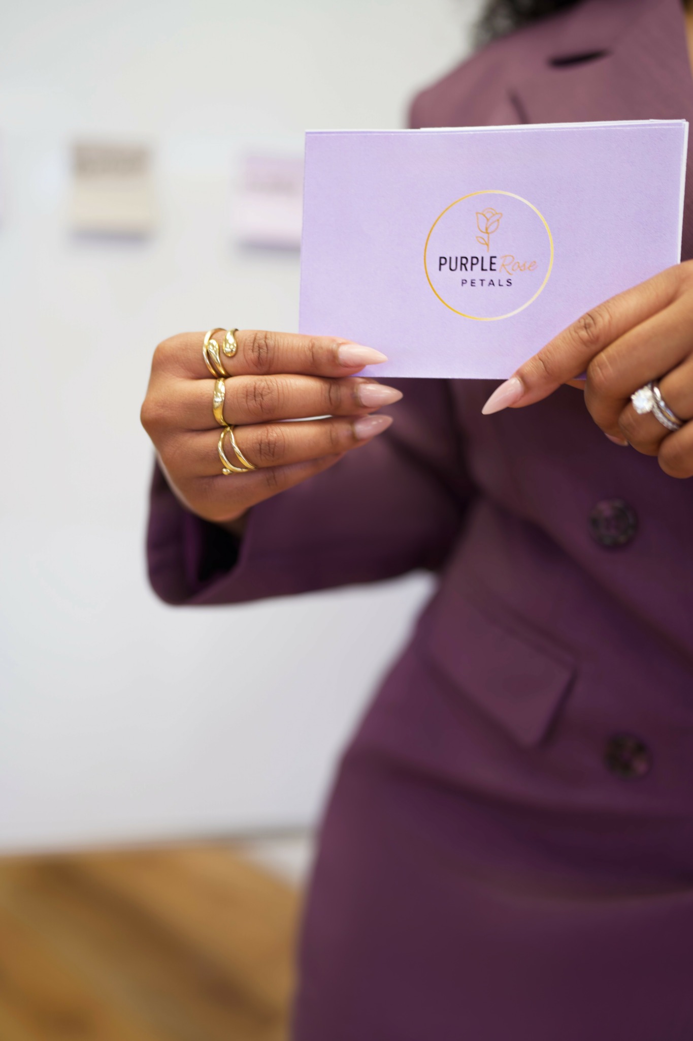 Person in purple blazer holding a purple card with gold lettering, rings on fingers, blurred background.