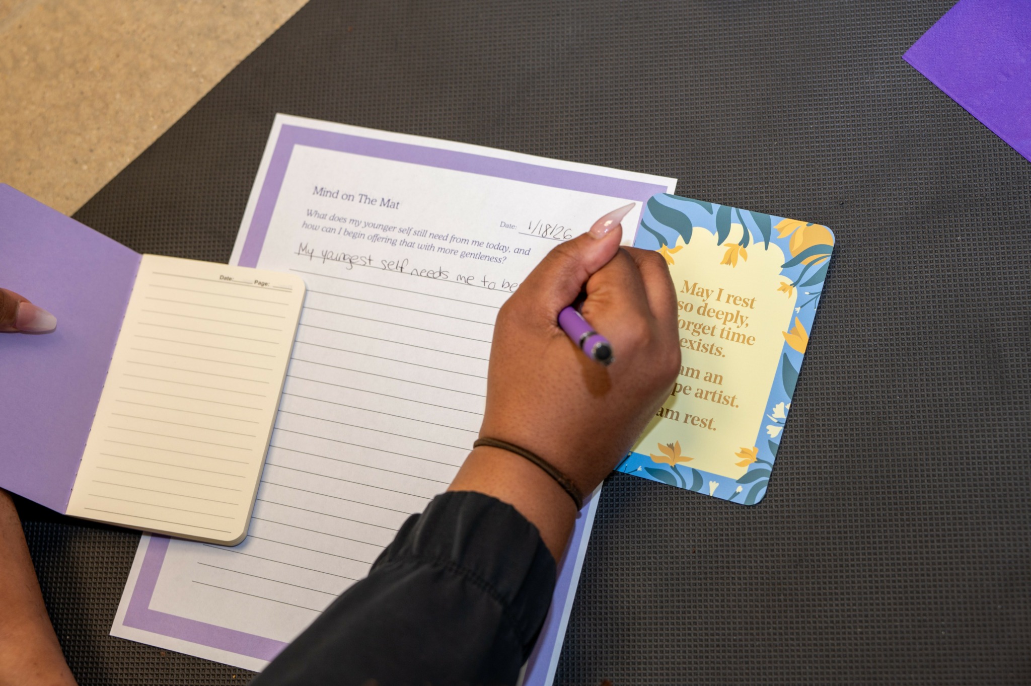 Person writing in a notebook with a purple pen, on a black surface with papers and a card nearby.