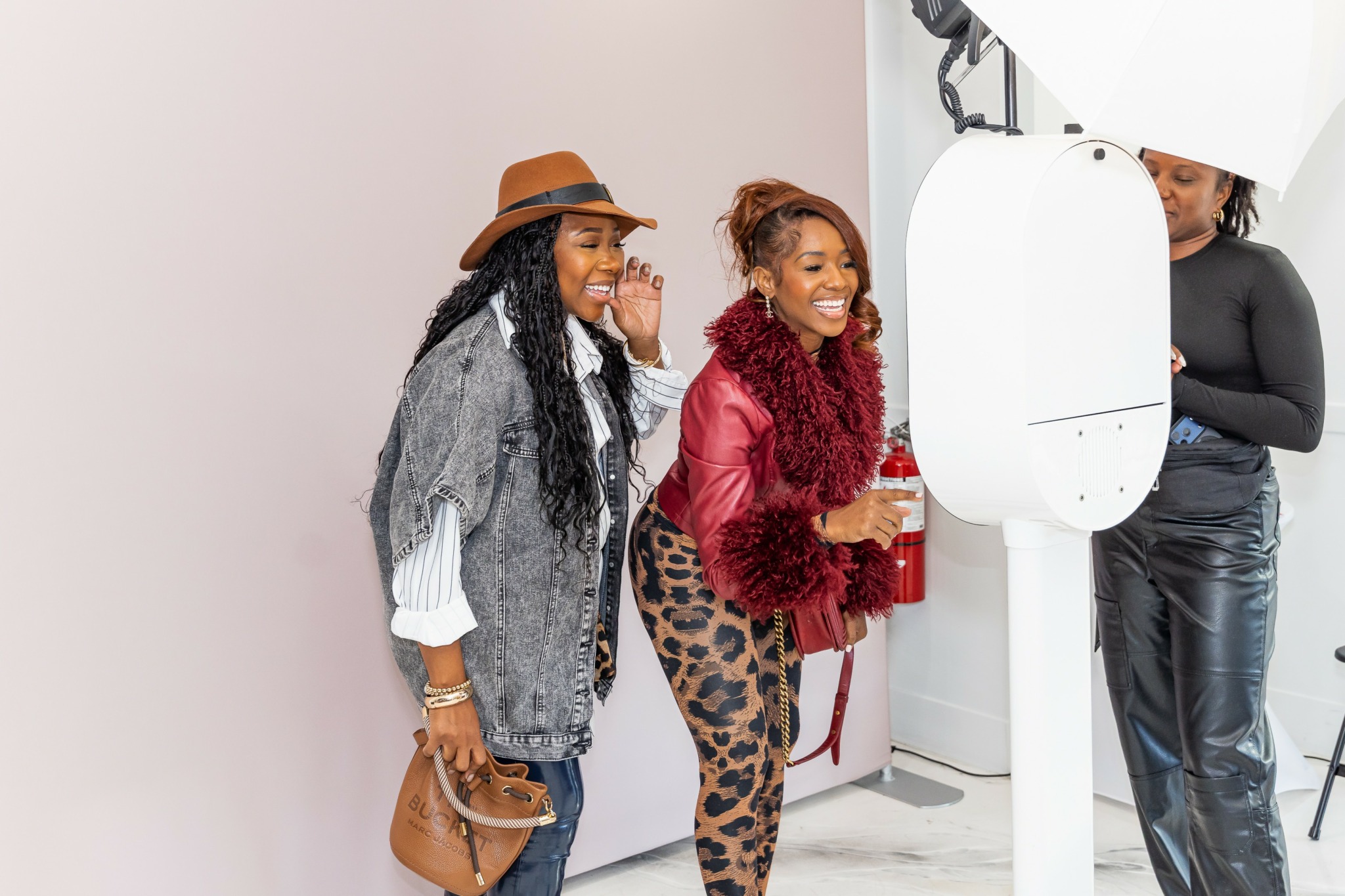 Two women smiling and talking in front of a photo booth, one wearing a hat and the other in a red jacket.