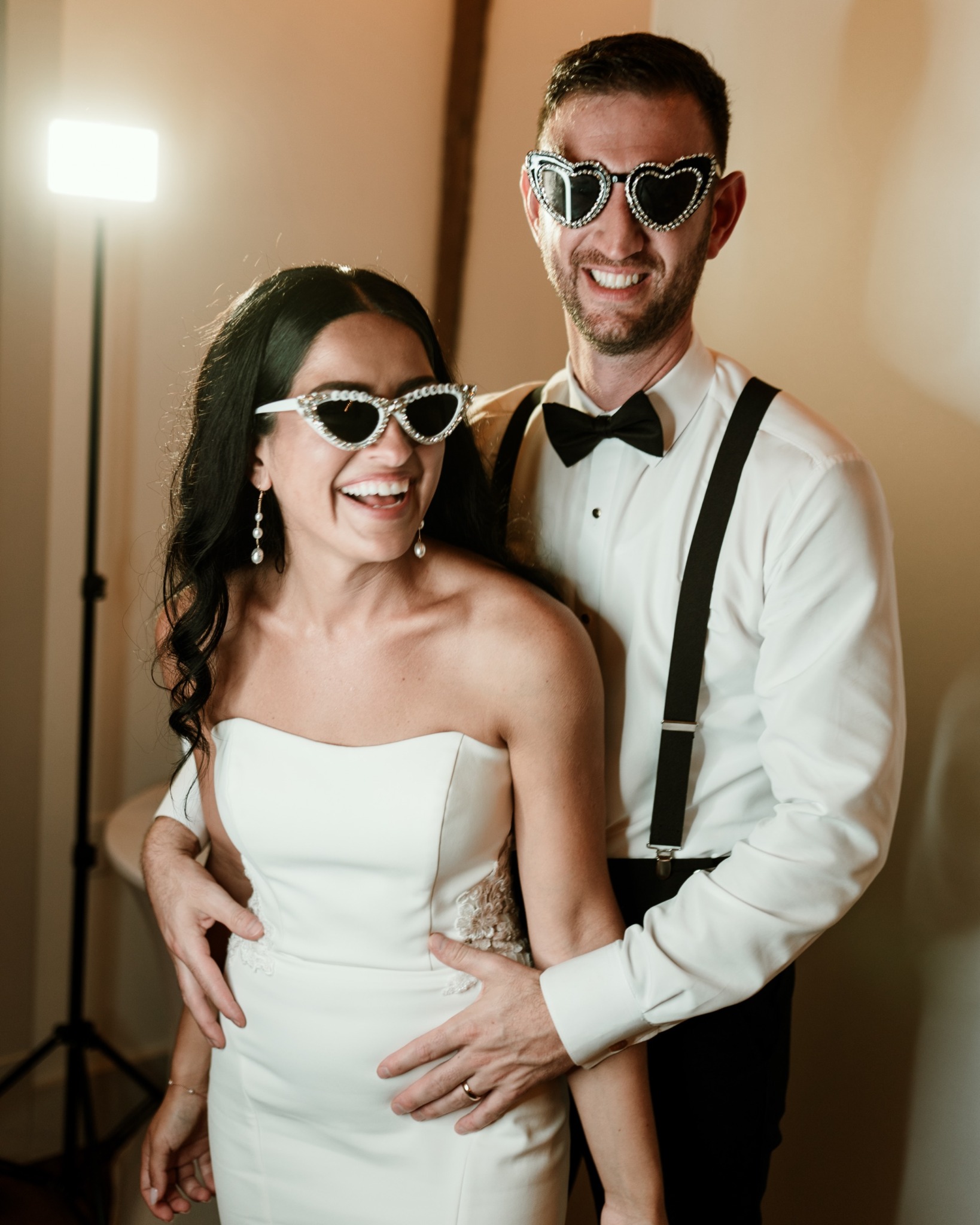 A smiling couple in formal attire wearing sunglasses, standing close together indoors with warm lighting.