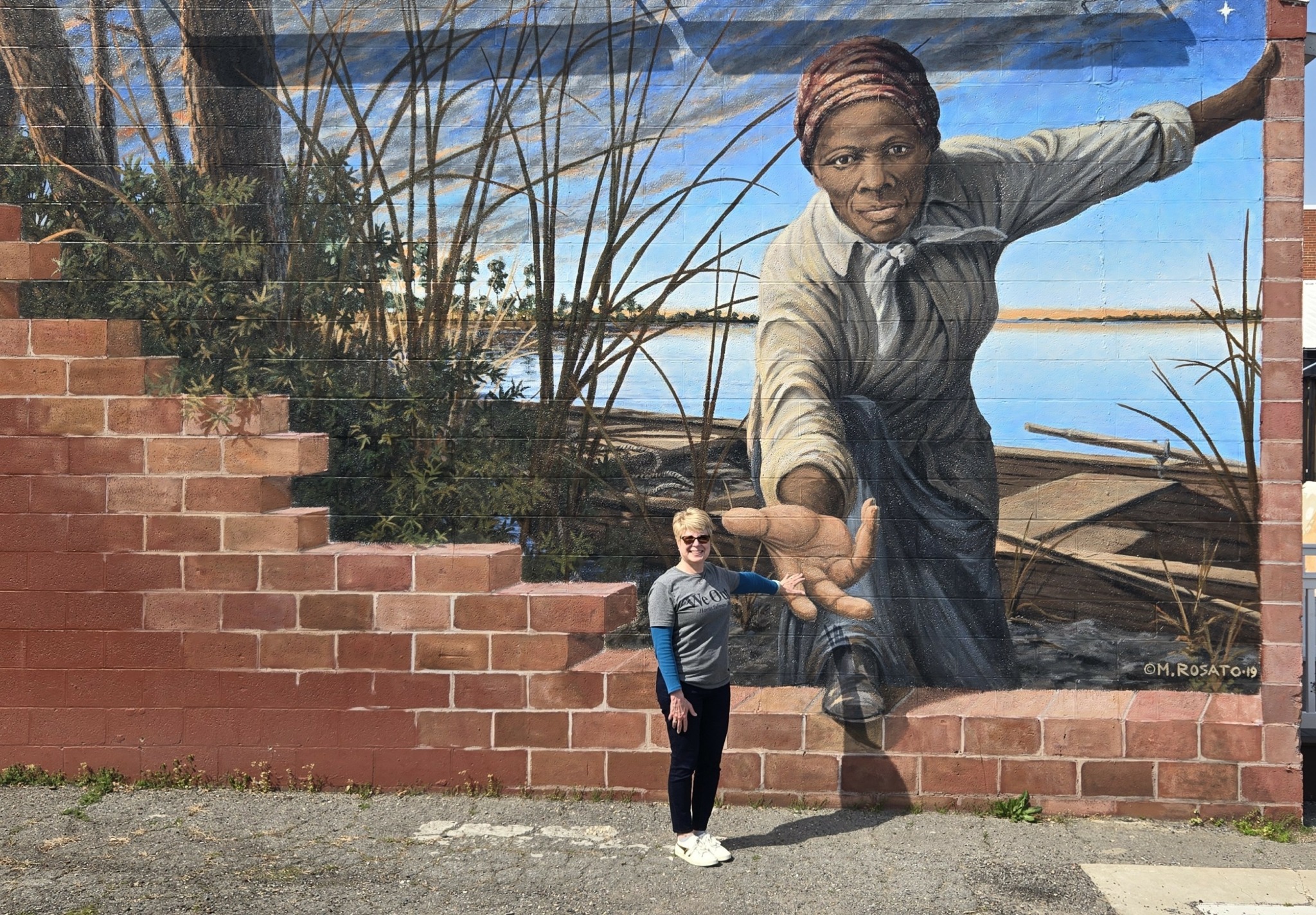 Person standing in front of a mural of a woman reaching out, with a brick wall and plants nearby.