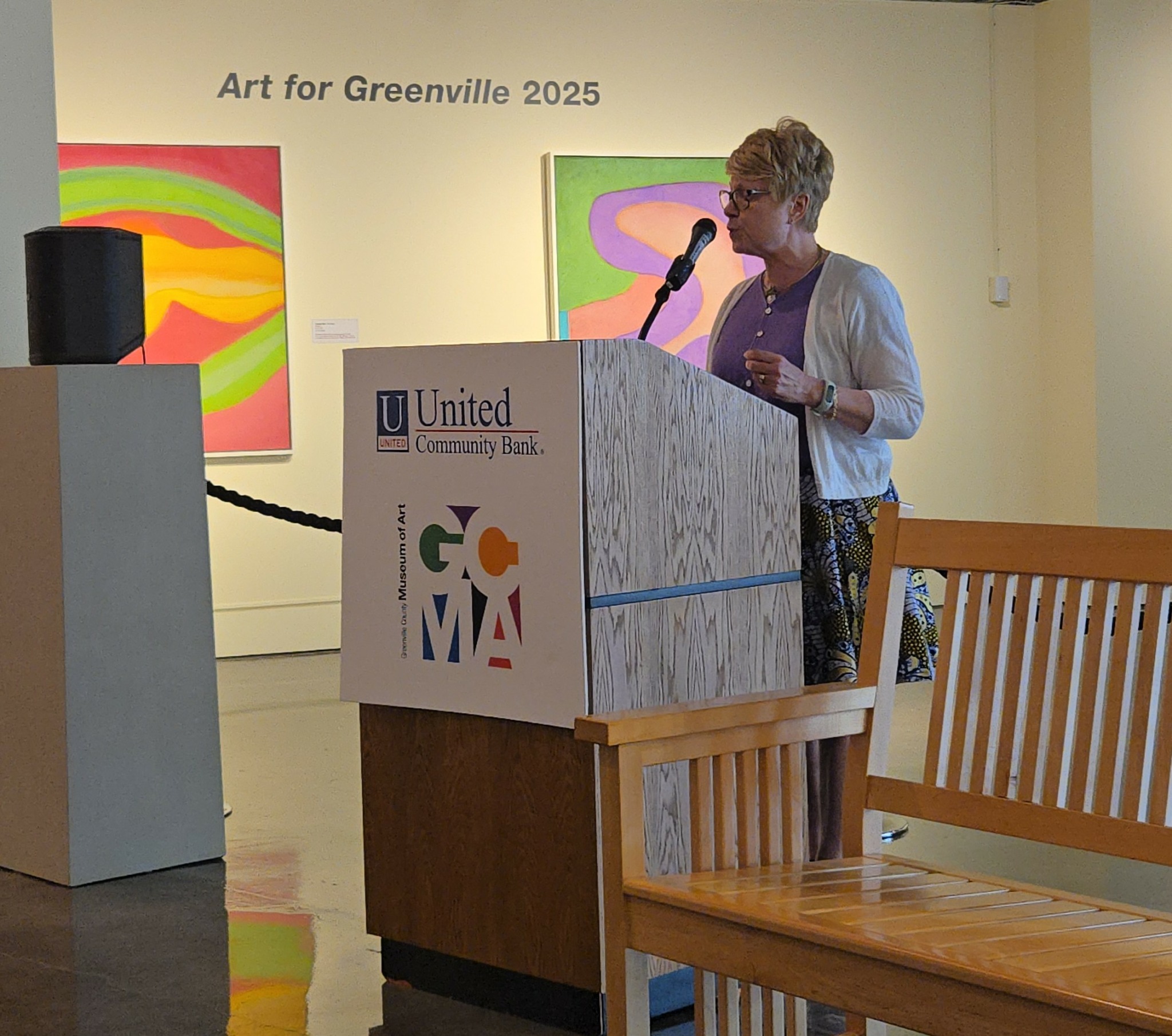 Woman speaking at a podium in an art gallery with colorful paintings on the wall.