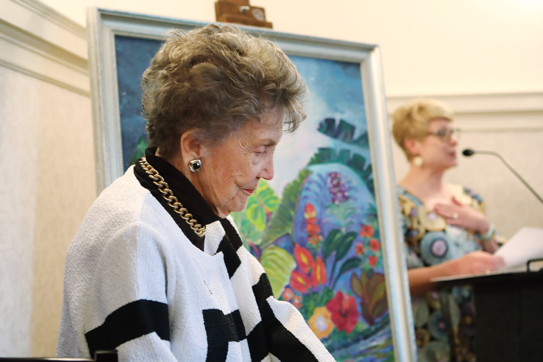 Older woman with short, curly hair, wearing a white and black striped sweater, earrings, and a gold chain necklace, standing near a colorful painting.