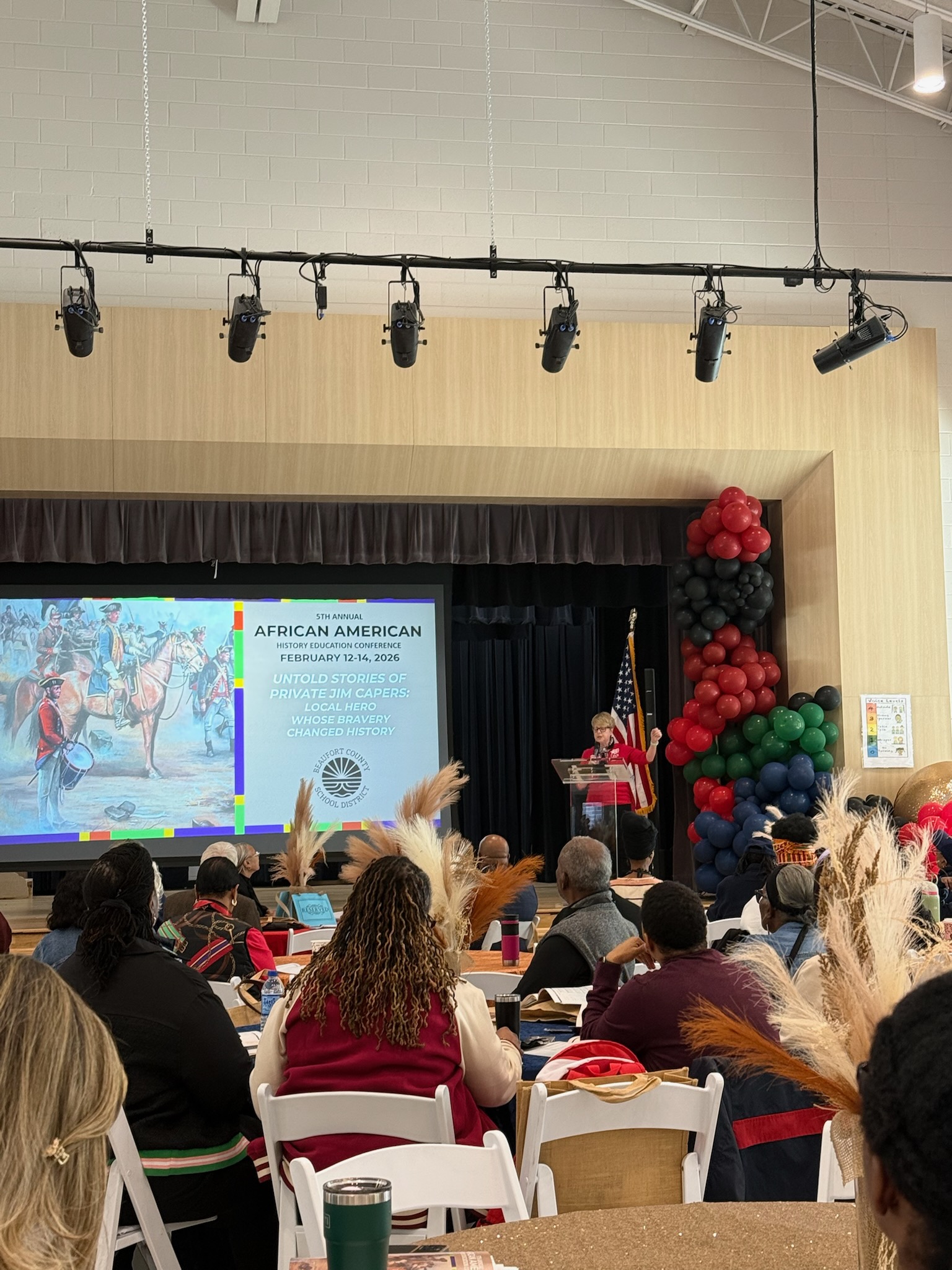 Audience seated in a hall watching a presentation with a large screen and colorful balloon decorations on the side.