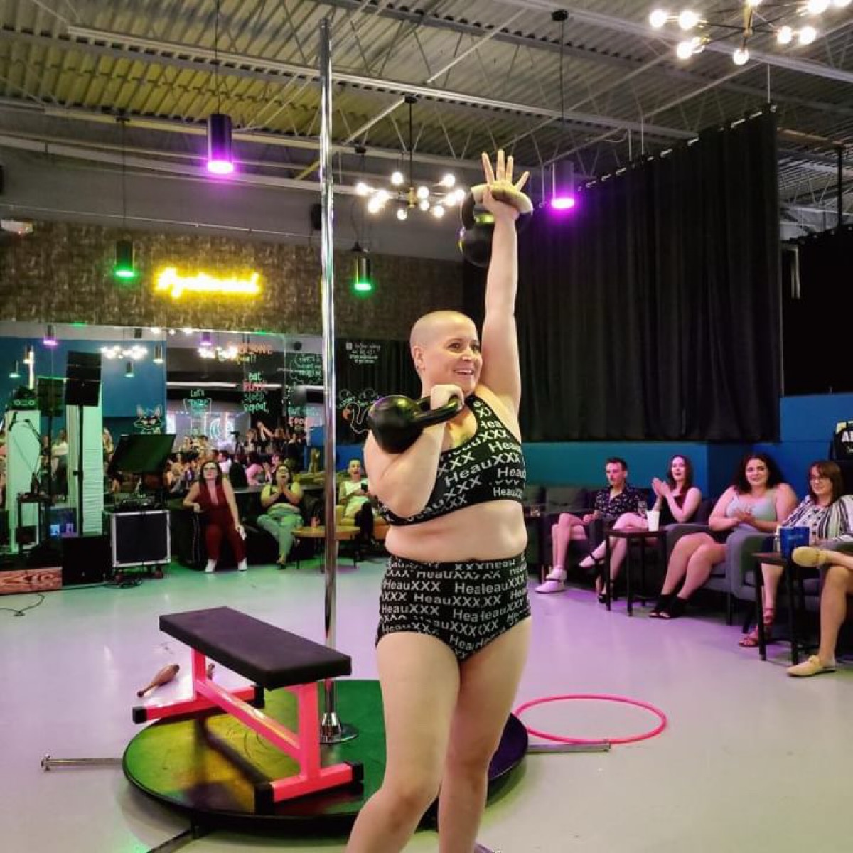 Performer with a bald head, wearing a black crop top and shorts, holding a kettlebell, raising one arm in a gym setting.
