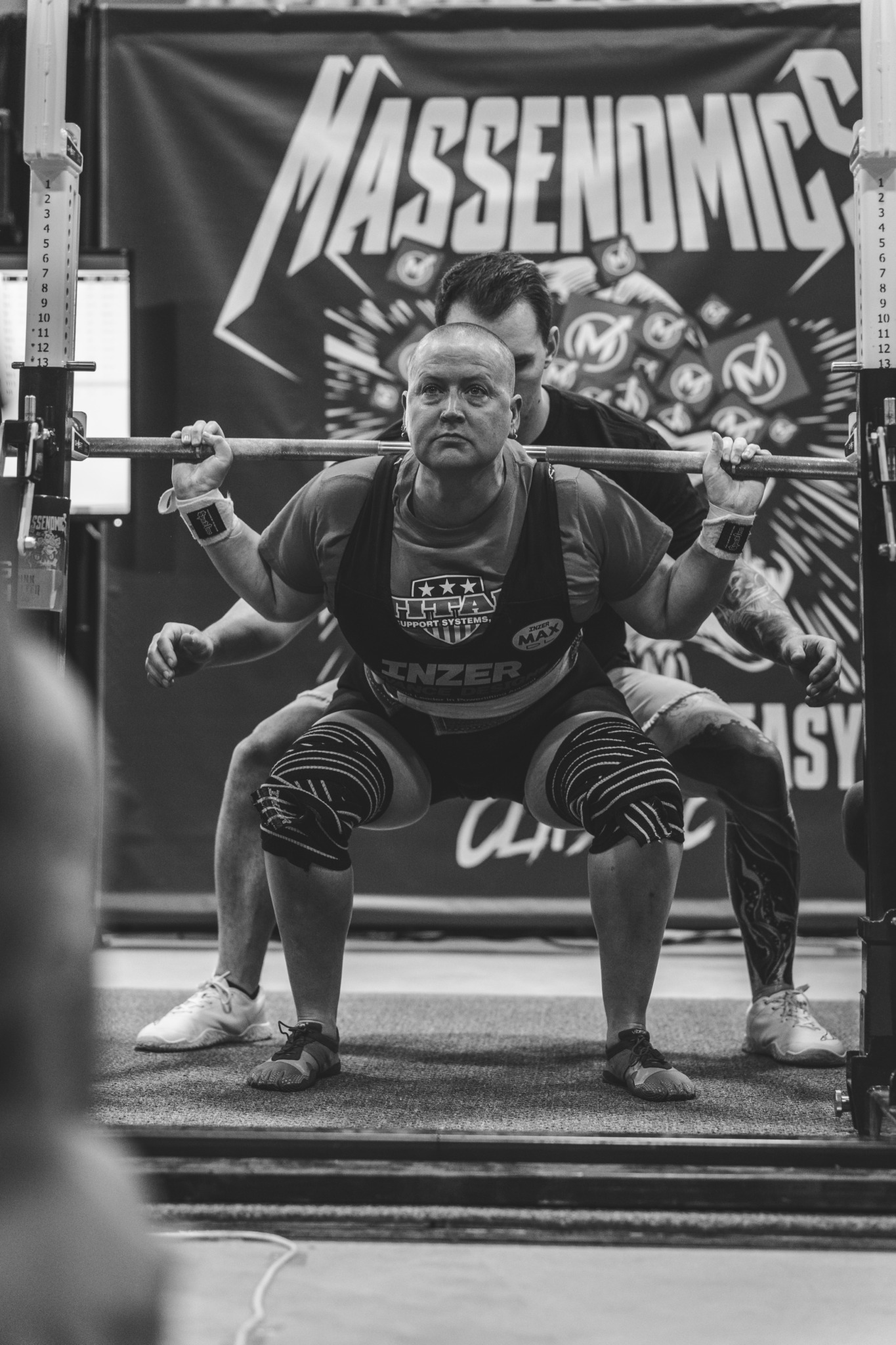 Person performing squat lift with barbell in gym, others in background, banner with 'Massonomics' logo behind.