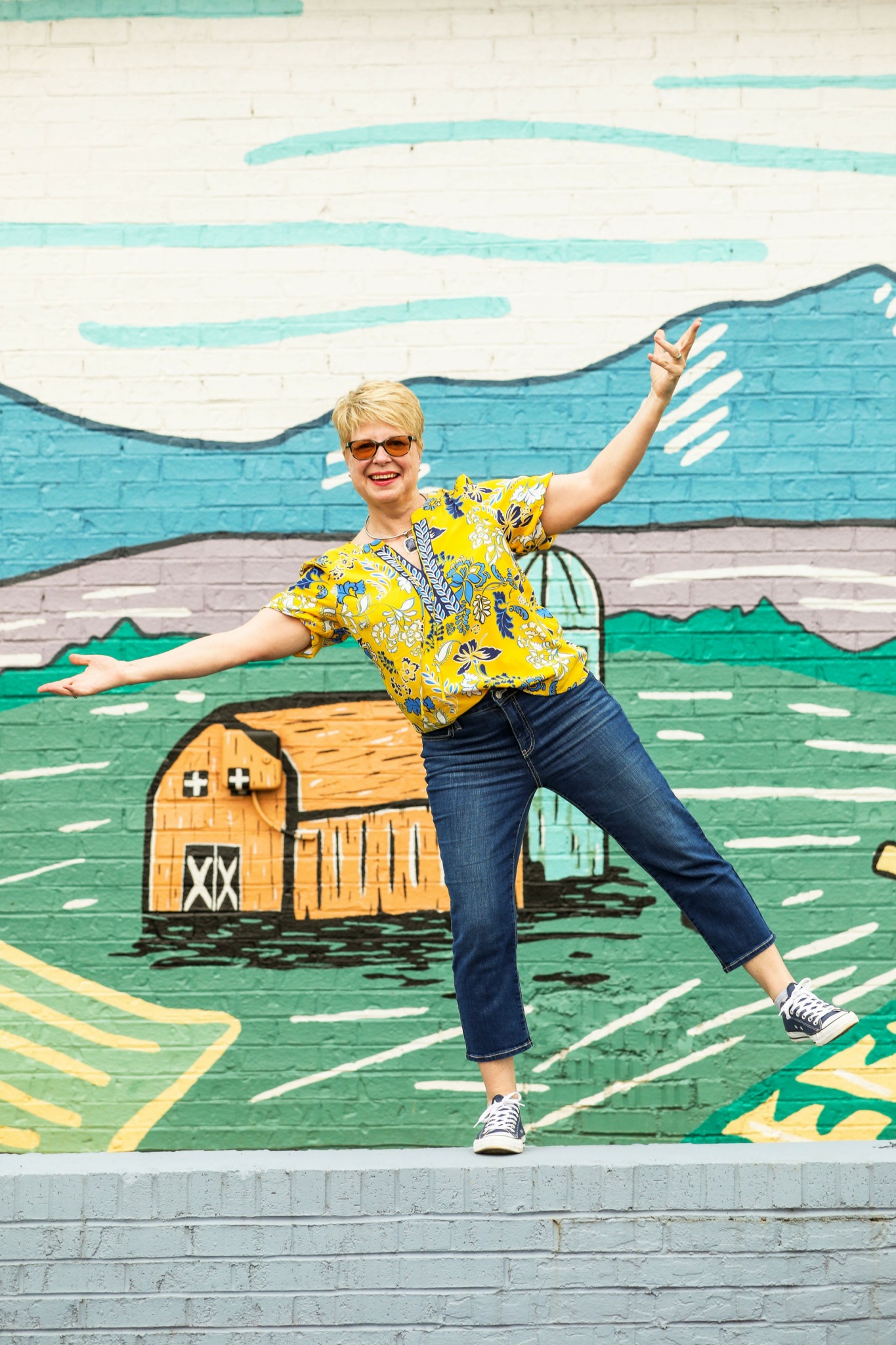 Person with short blonde hair and glasses stands in front of a colorful mural, arms outstretched, smiling.