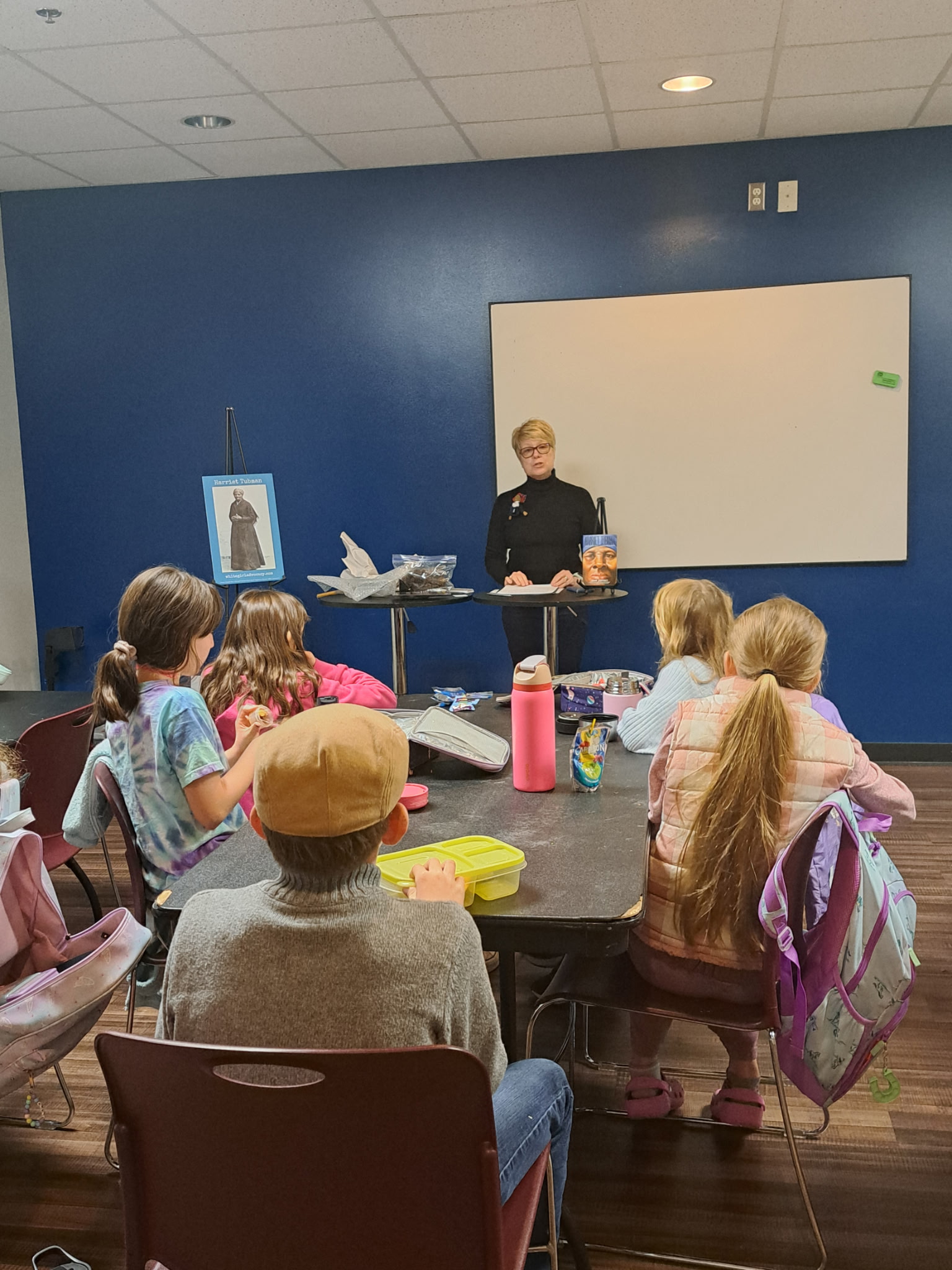 Teacher standing at front of classroom with students seated at tables, whiteboard behind, and a portrait on the wall.