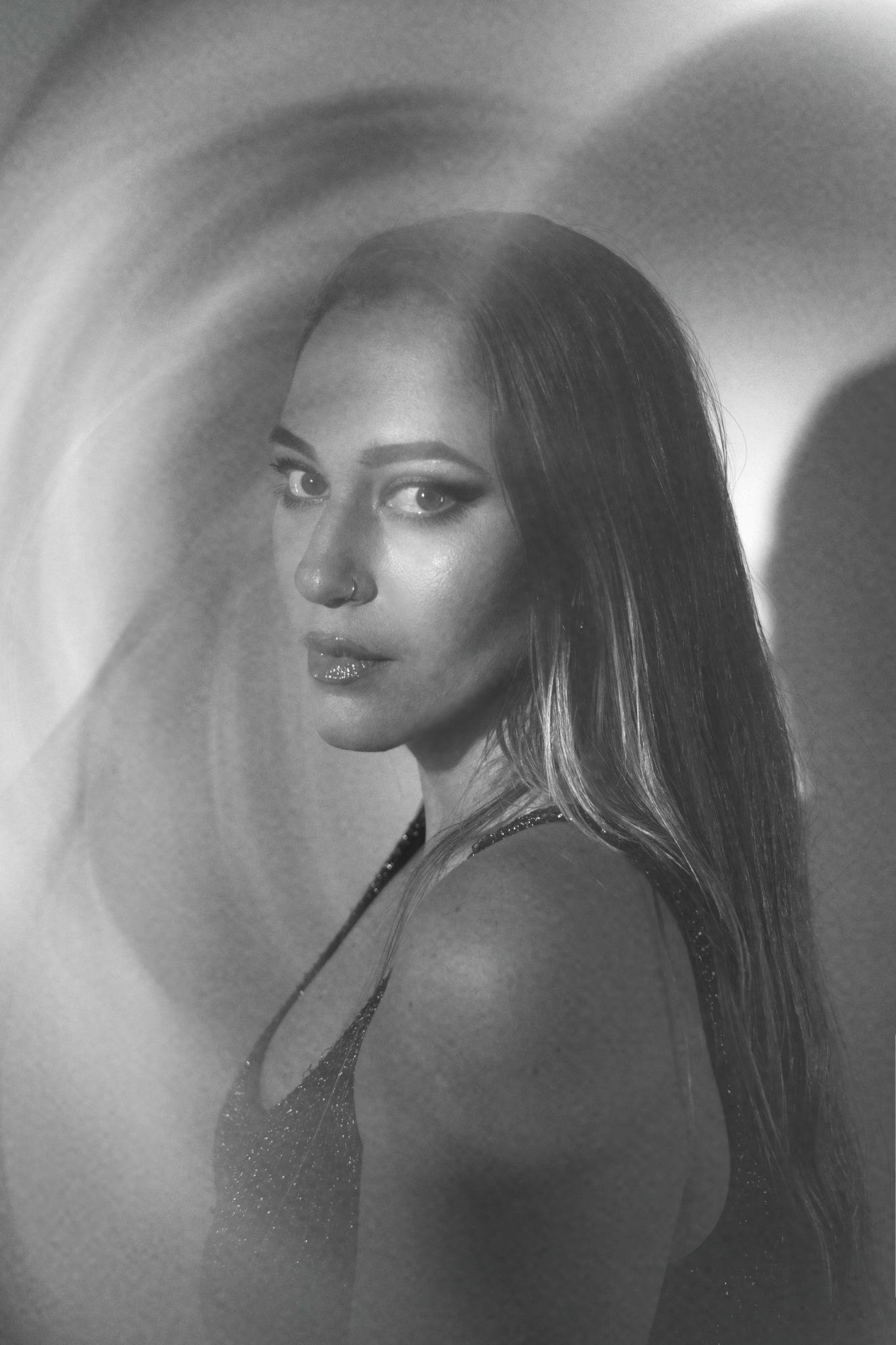 A woman with long hair looking at the camera, in black and white, with a blurred background.