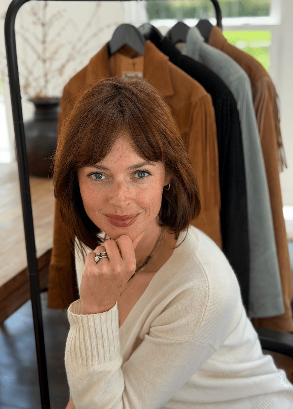 Woman with brown hair and blue eyes, wearing a white sweater, standing near a clothing rack.