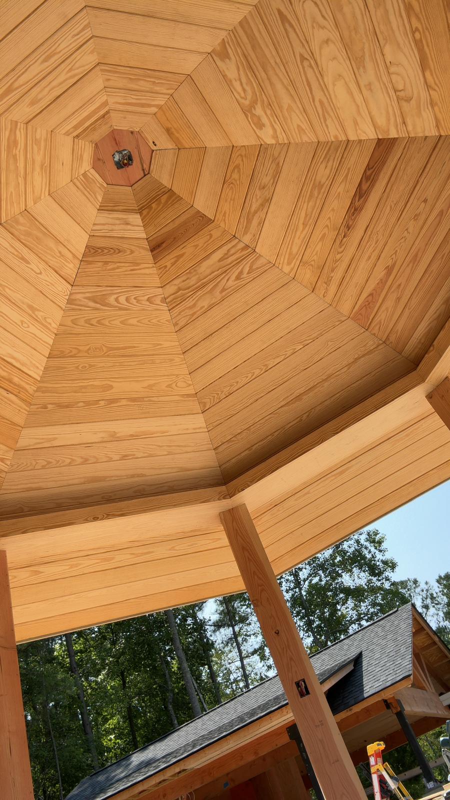 Looking up at a wooden ceiling with a central vent, surrounded by wooden panels, with trees and sky visible outside.