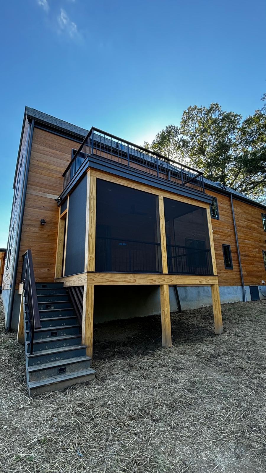 Elevated wooden deck with black screen panels, stairs on left, attached to a wooden house with trees in background.