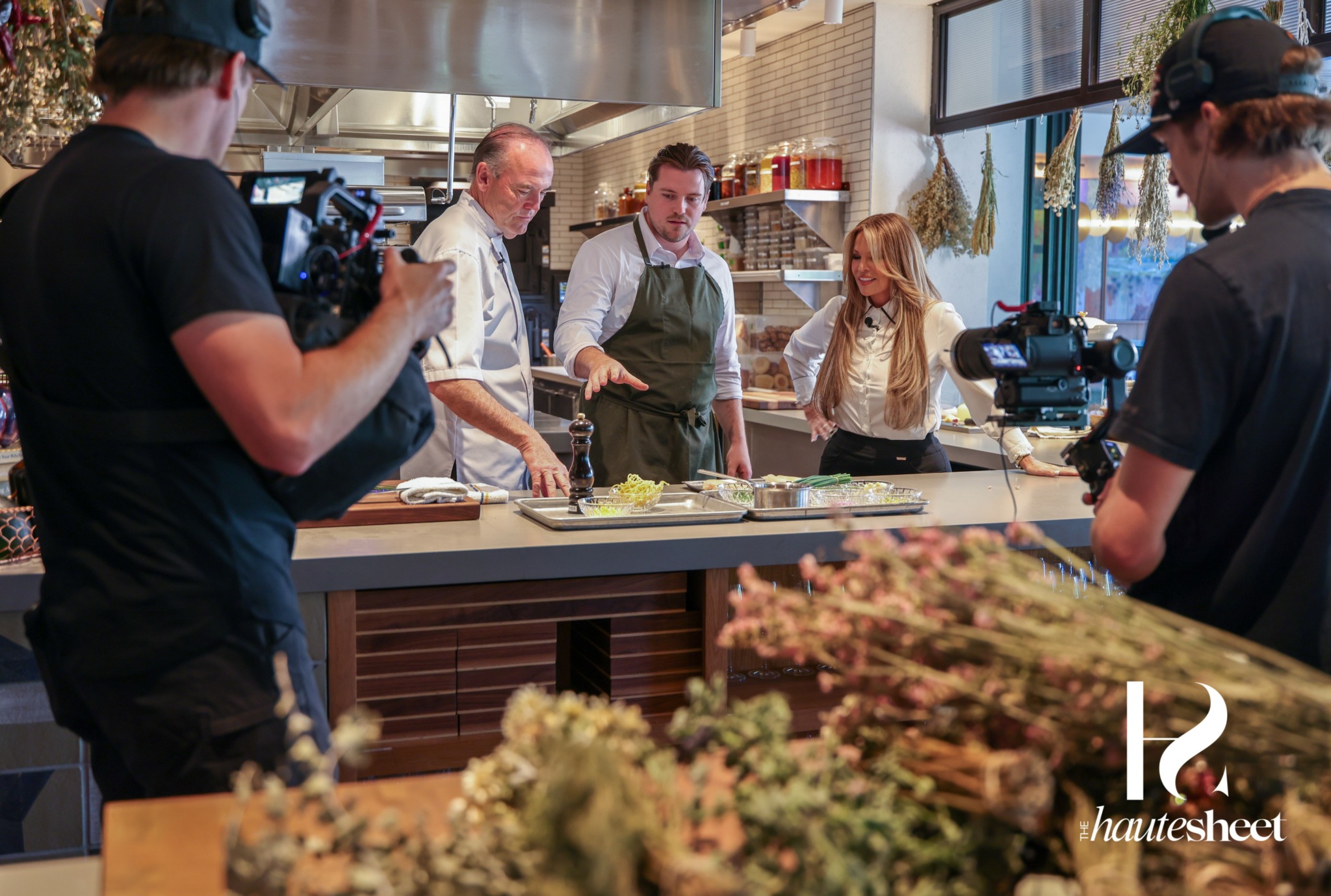 Four people in a kitchen, two chefs and two customers, with cameras filming the scene, flowers in foreground.