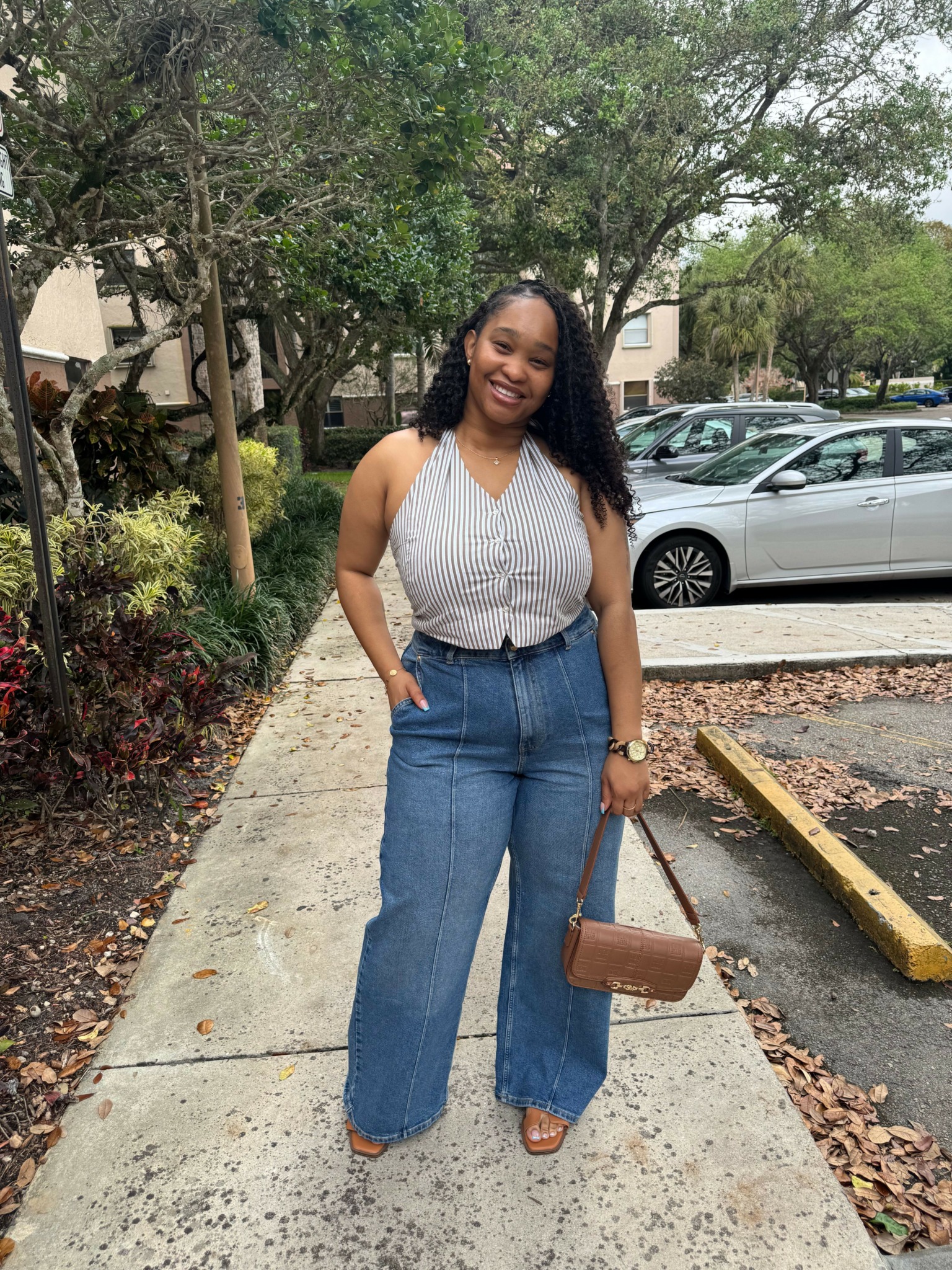 Woman with long dark hair in white top and blue jeans standing on sidewalk near trees and parked cars.