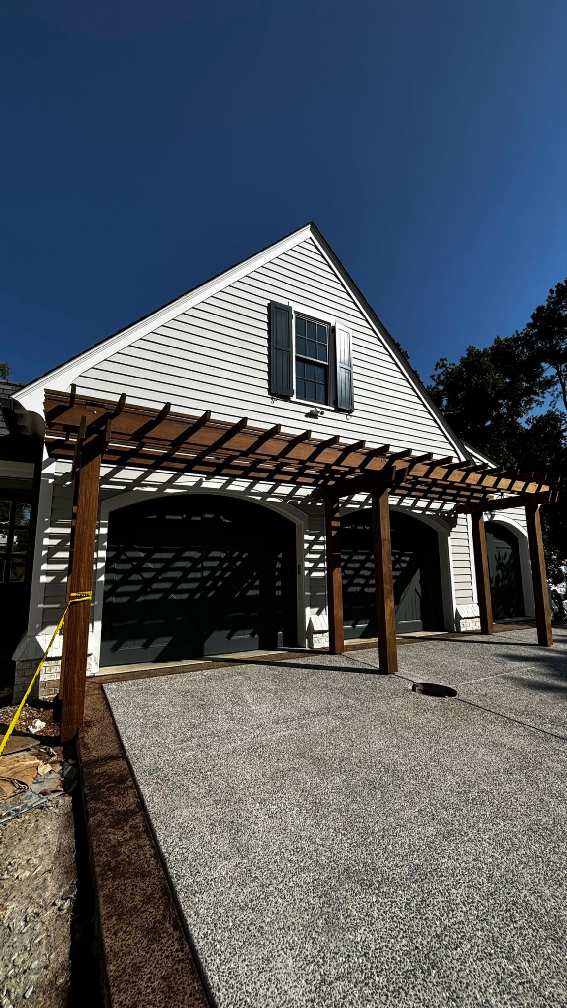 White house with black shutters, wooden pergola, and driveway, under a clear blue sky.