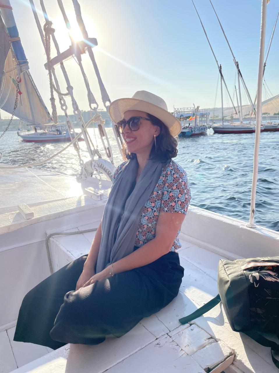 Woman wearing a hat and sunglasses sitting on a boat with water and sailboats in background.