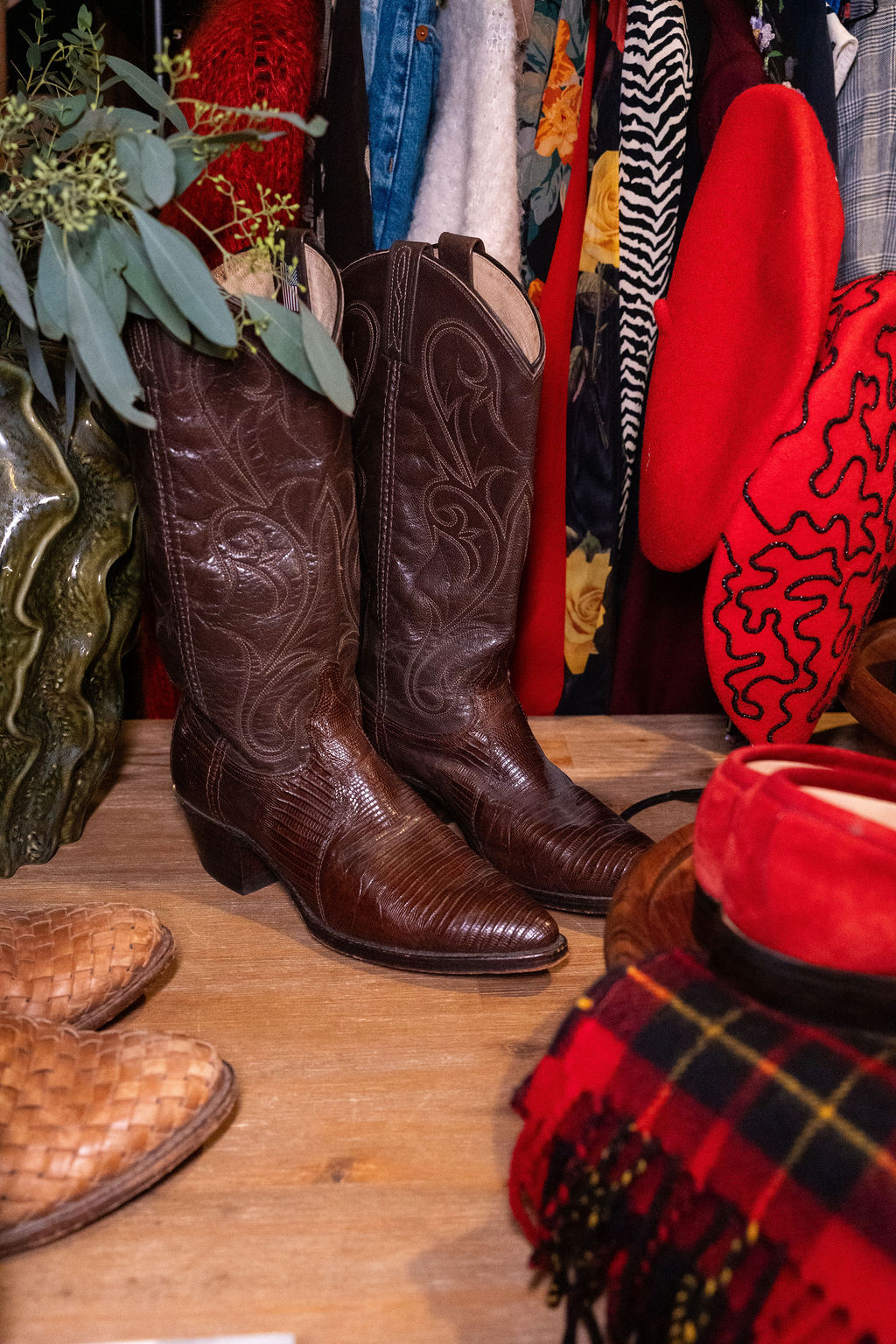 Pair of brown cowboy boots on a wooden surface with clothing and accessories in background.