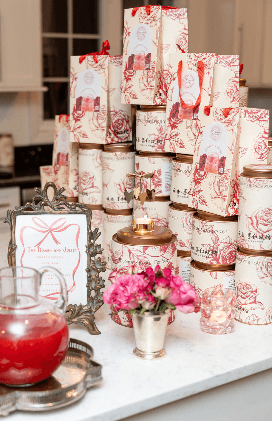 Stacked gift boxes with red and white patterns, pink flowers in a silver pot, and a framed sign on a table.