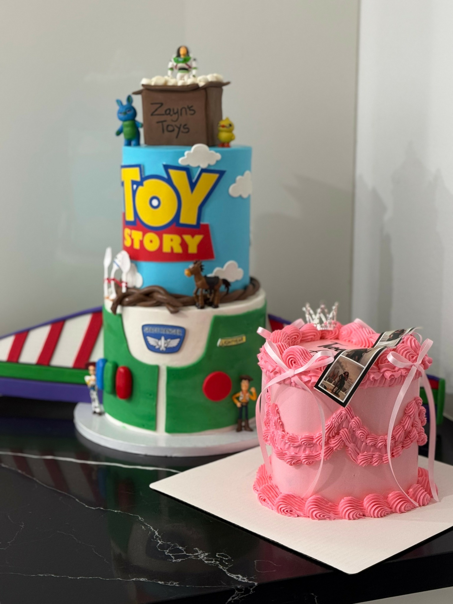 Two decorated birthday cakes, one themed with Toy Story characters and the other pink with a photo frame, on a black surface.