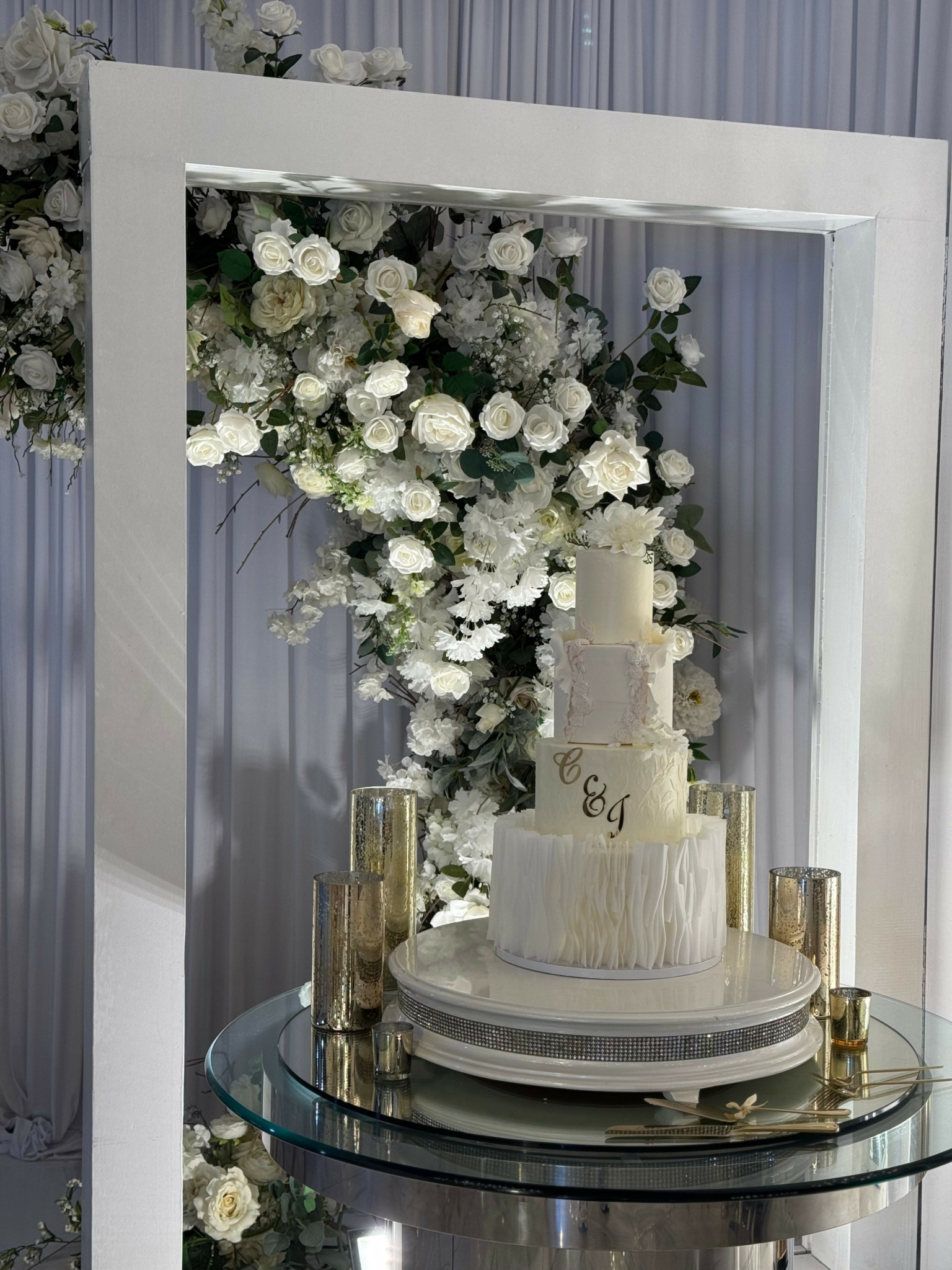 Wedding cake with white flowers and gold accents on a glass table, framed by a white rectangular structure.