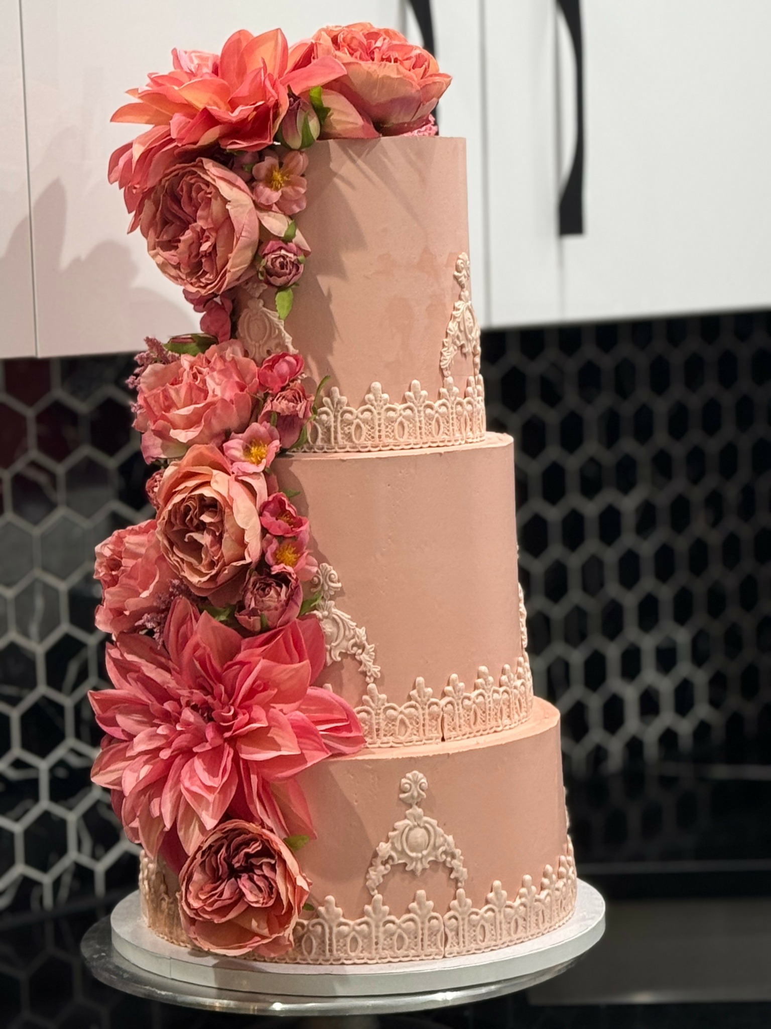 Three-tiered pink cake decorated with pink flowers and intricate white icing patterns, on a white cake stand.