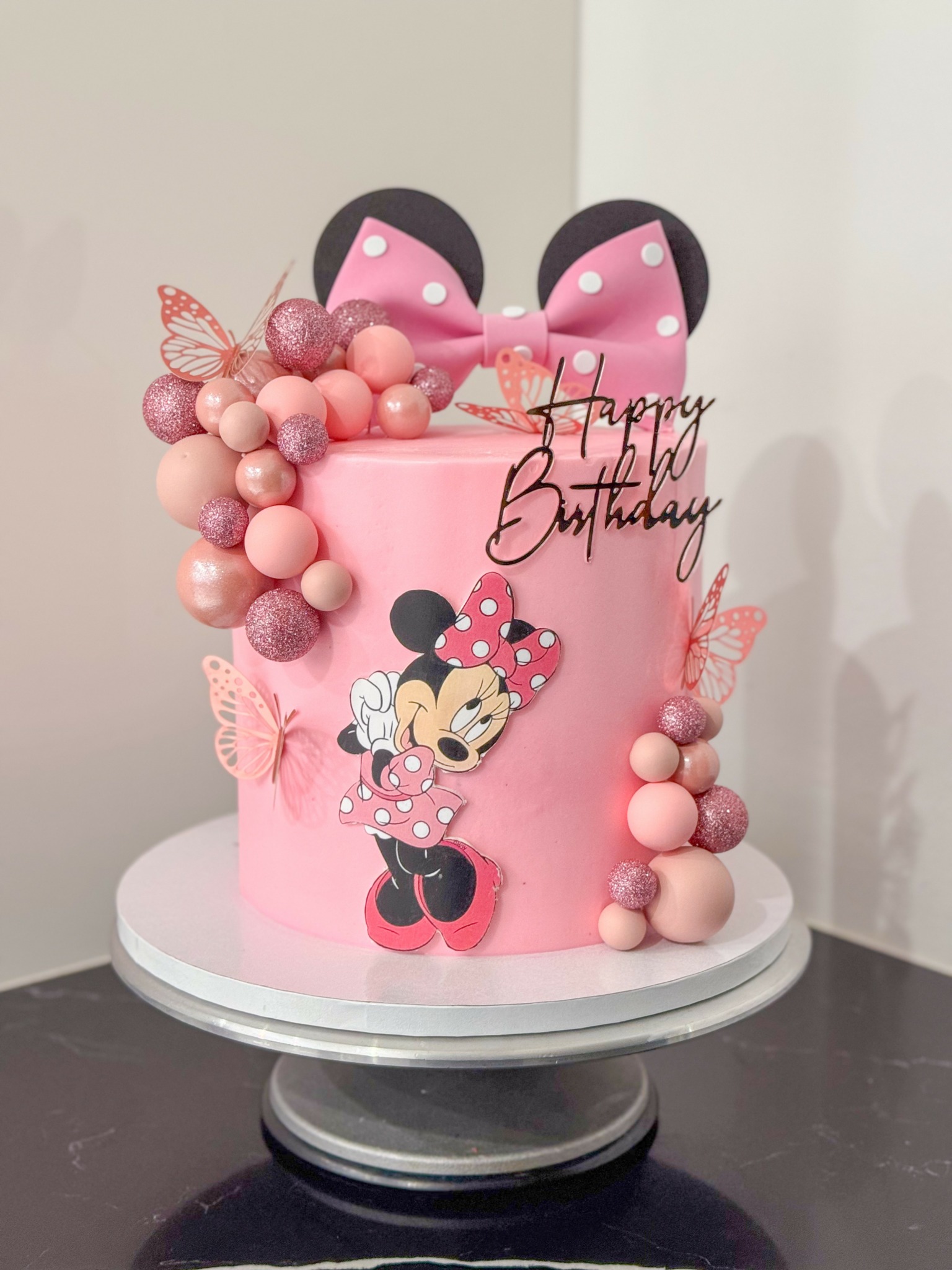 Pink birthday cake with Minnie Mouse decoration, pink bows, and pink spherical accents, on a white cake stand.