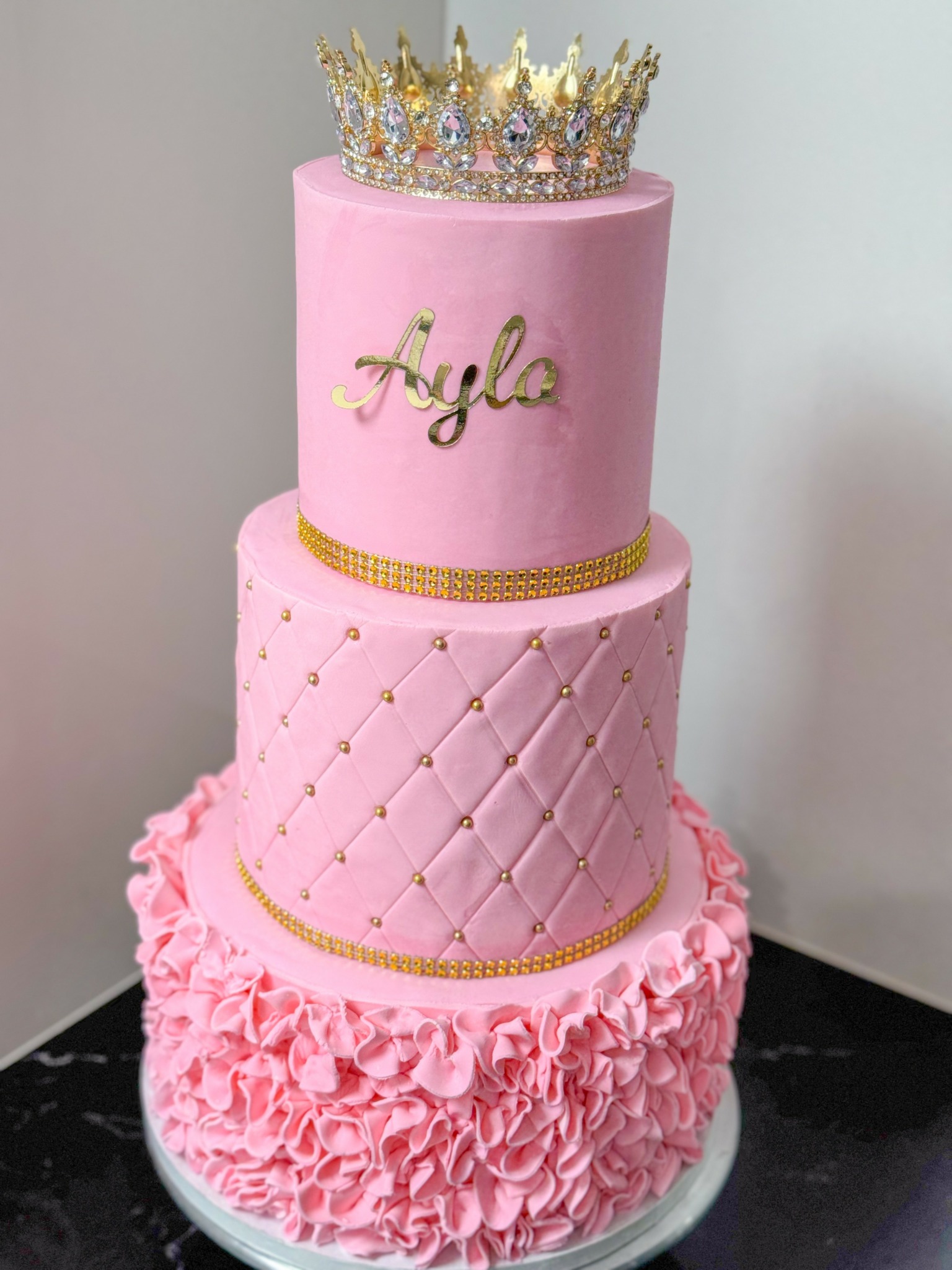 Three-tier pink cake with gold accents, a crown on top, and ruffled decoration at the bottom.