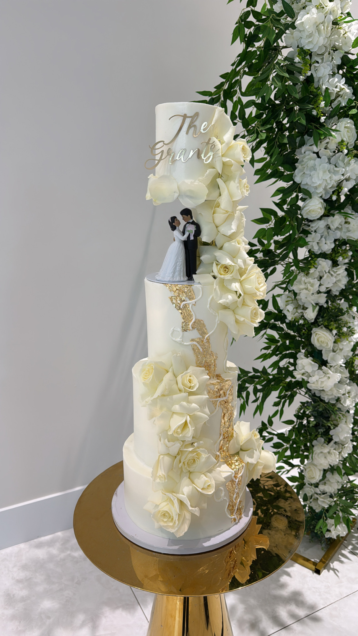 Tall wedding cake with white flowers and a bride and groom topper, on a gold stand next to floral arrangements.