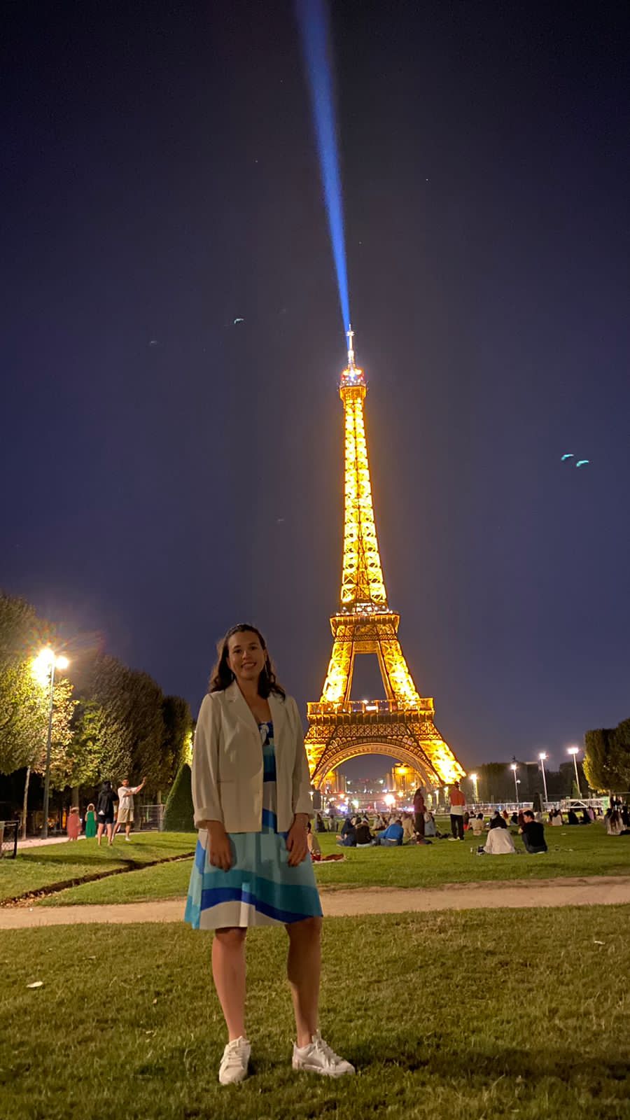 Person standing on grass in front of illuminated Eiffel Tower at night, with blue beam of light extending upward.