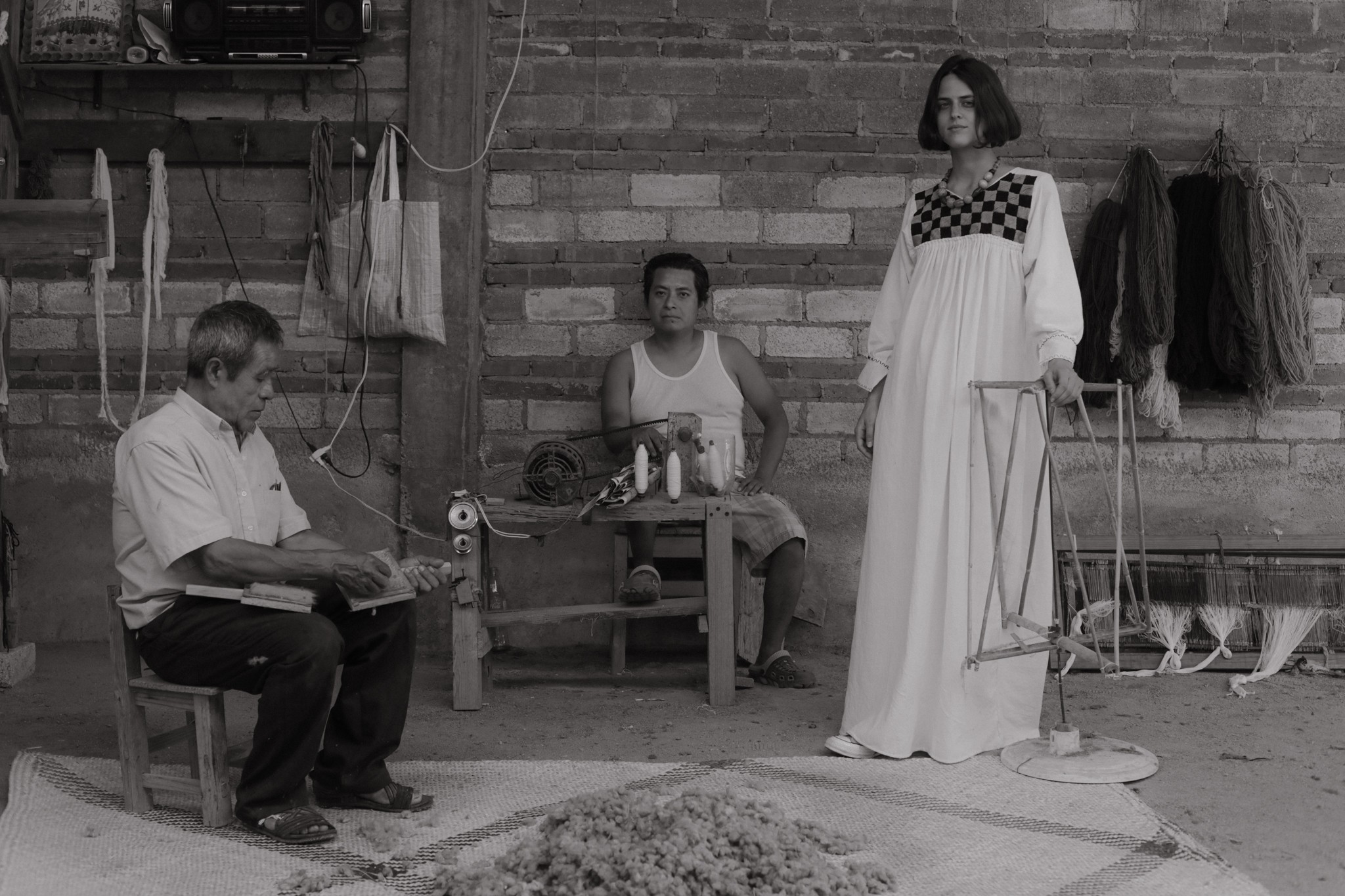 Three people in a room with a brick wall; one seated reading, one seated behind a table, one standing with a dress.