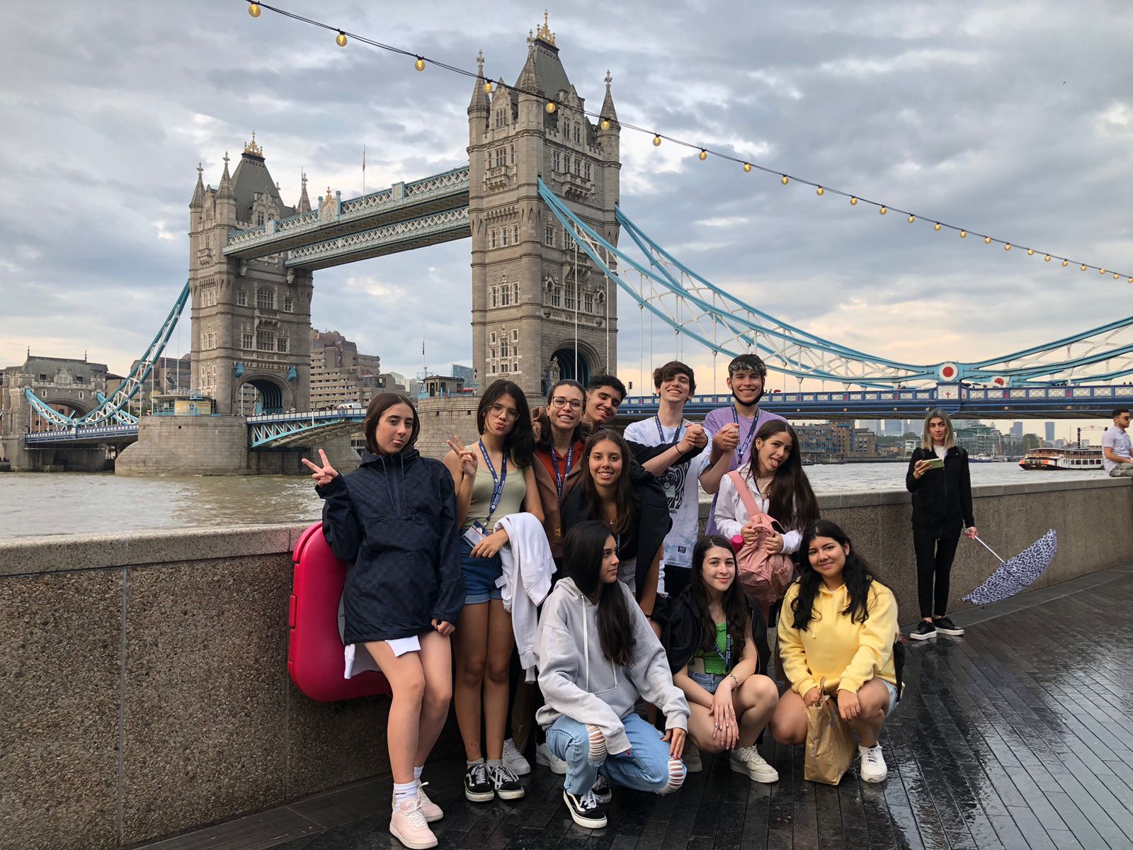 Group of people posing in front of Tower Bridge in London, some making peace signs, on a cloudy day.