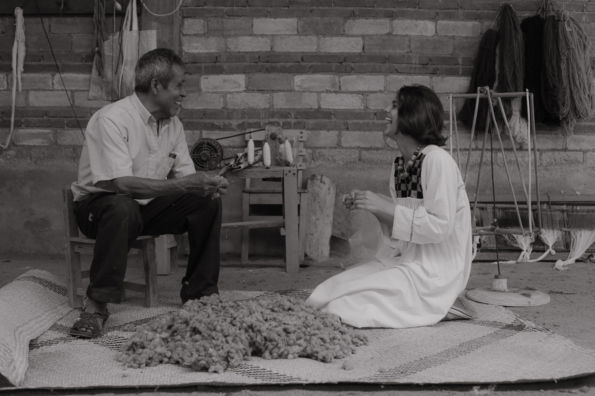 A man and a woman sit on a mat in front of a brick wall, engaging in conversation or activity.