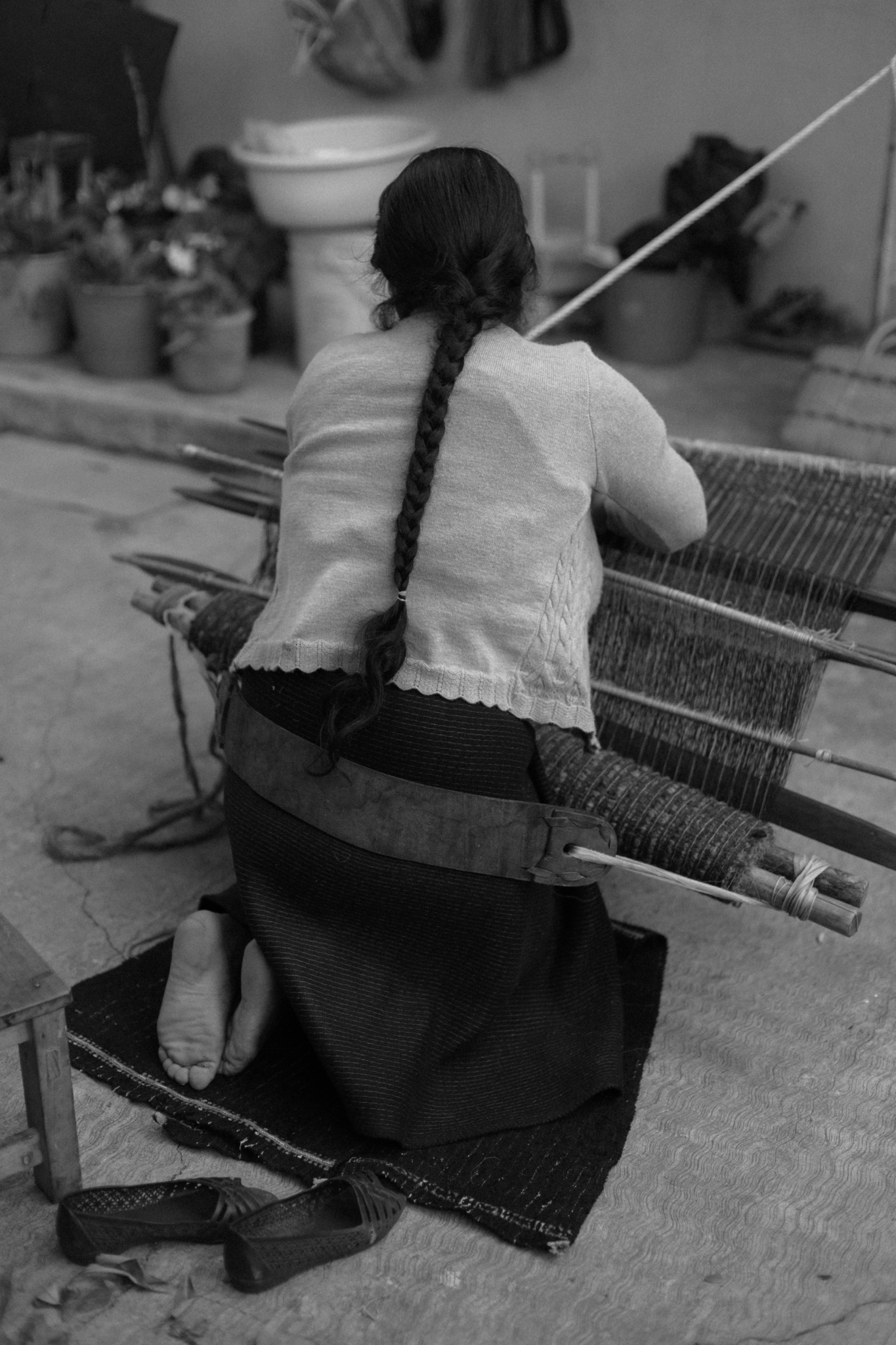 Person with long braided hair sitting on the ground, weaving or working with a loom or similar equipment.