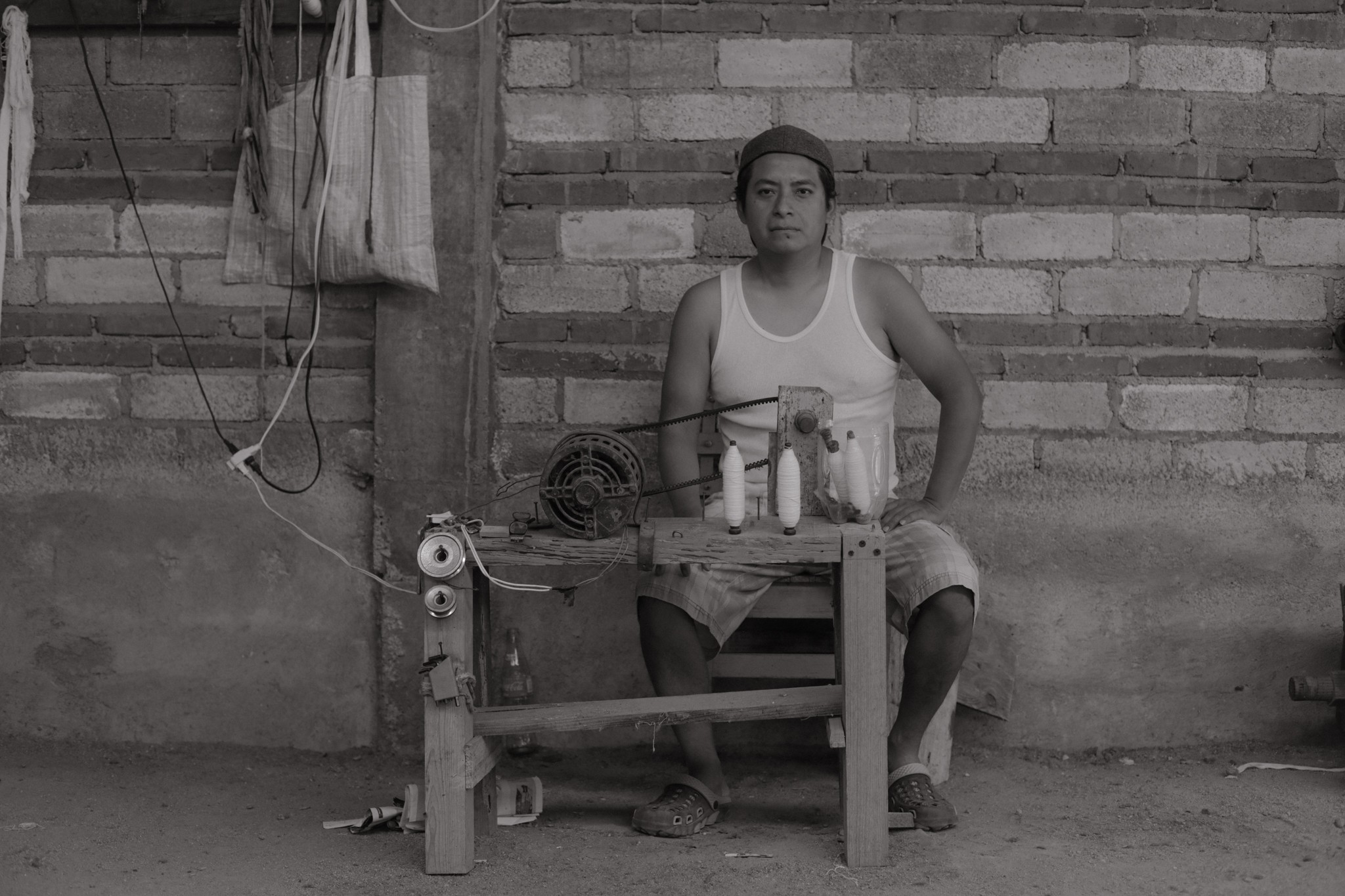 Young person sitting on a wooden bench in front of a brick wall, with a table and objects nearby.