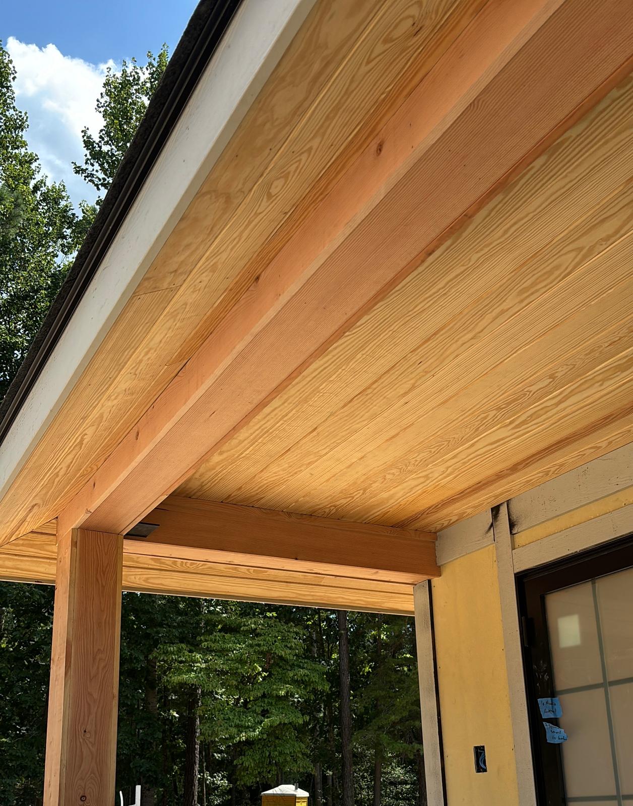 Close-up of wooden roof overhang with visible wood grain and support beams, with trees and sky in background.
